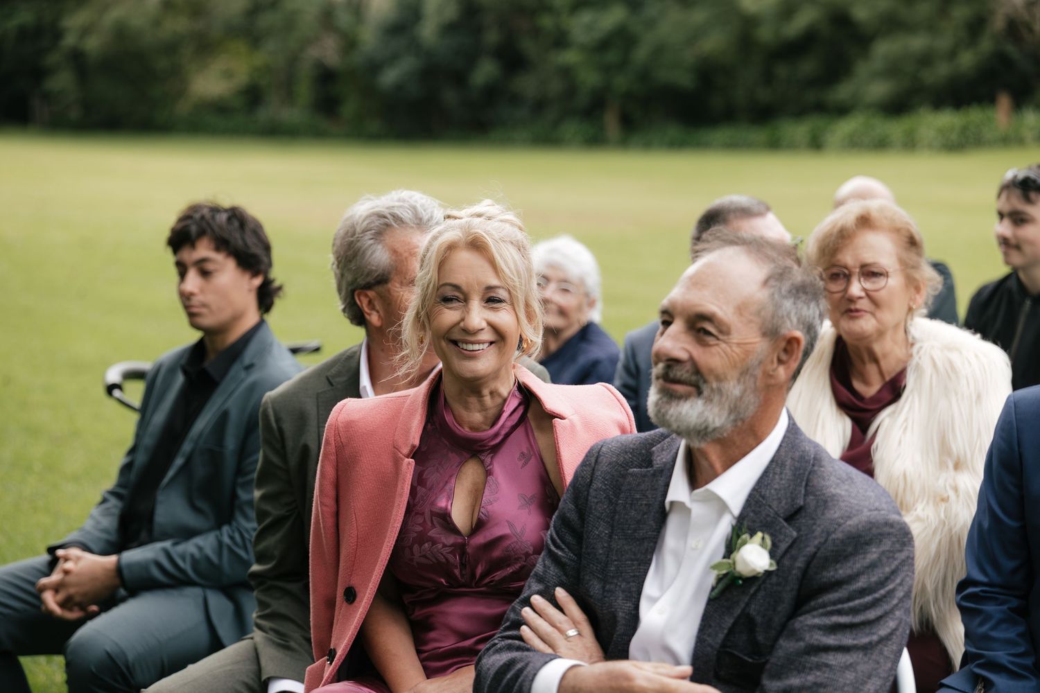 Guests smiling and enjoying an outdoor wedding ceremony seated on chairs in a lush green garden setting.