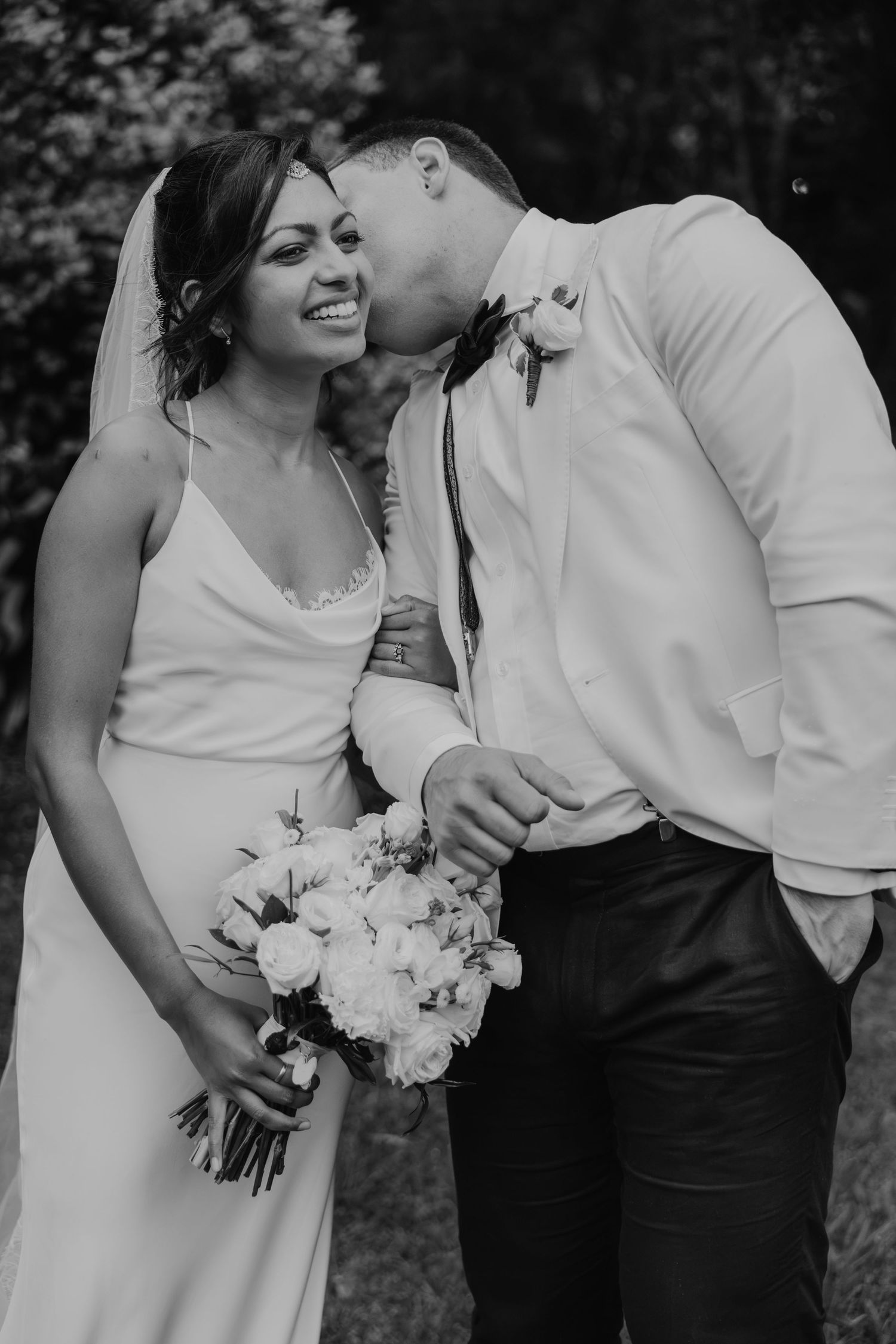 Black and white photo of a groom kissing his bride on the cheek as she smiles holding a white floral bouquet.