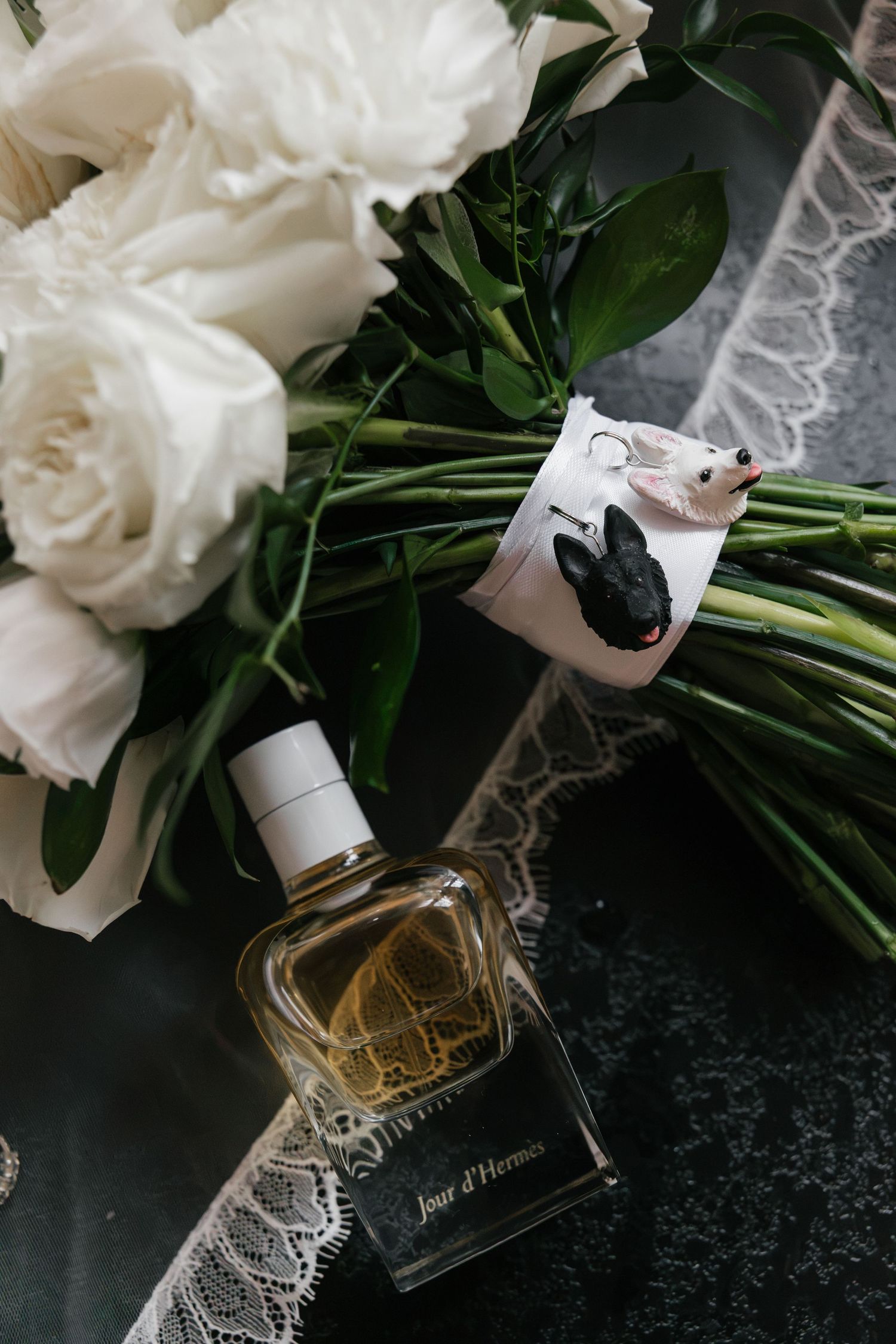 Jour d'Hermes perfume bottle beside white roses bouquet with green stems and lace on dark surface.