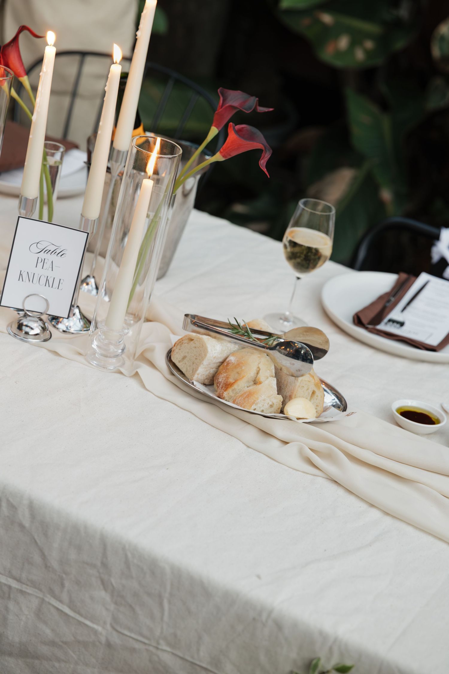 Elegant wedding table setting with white linen, candles, calla lilies, bread rolls, and a glass of white wine.