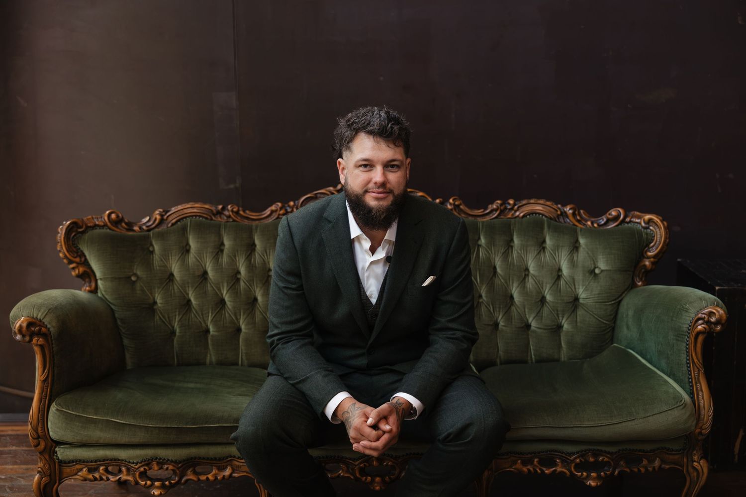 Man in dark suit sitting on ornate green velvet sofa against moody brown backdrop.