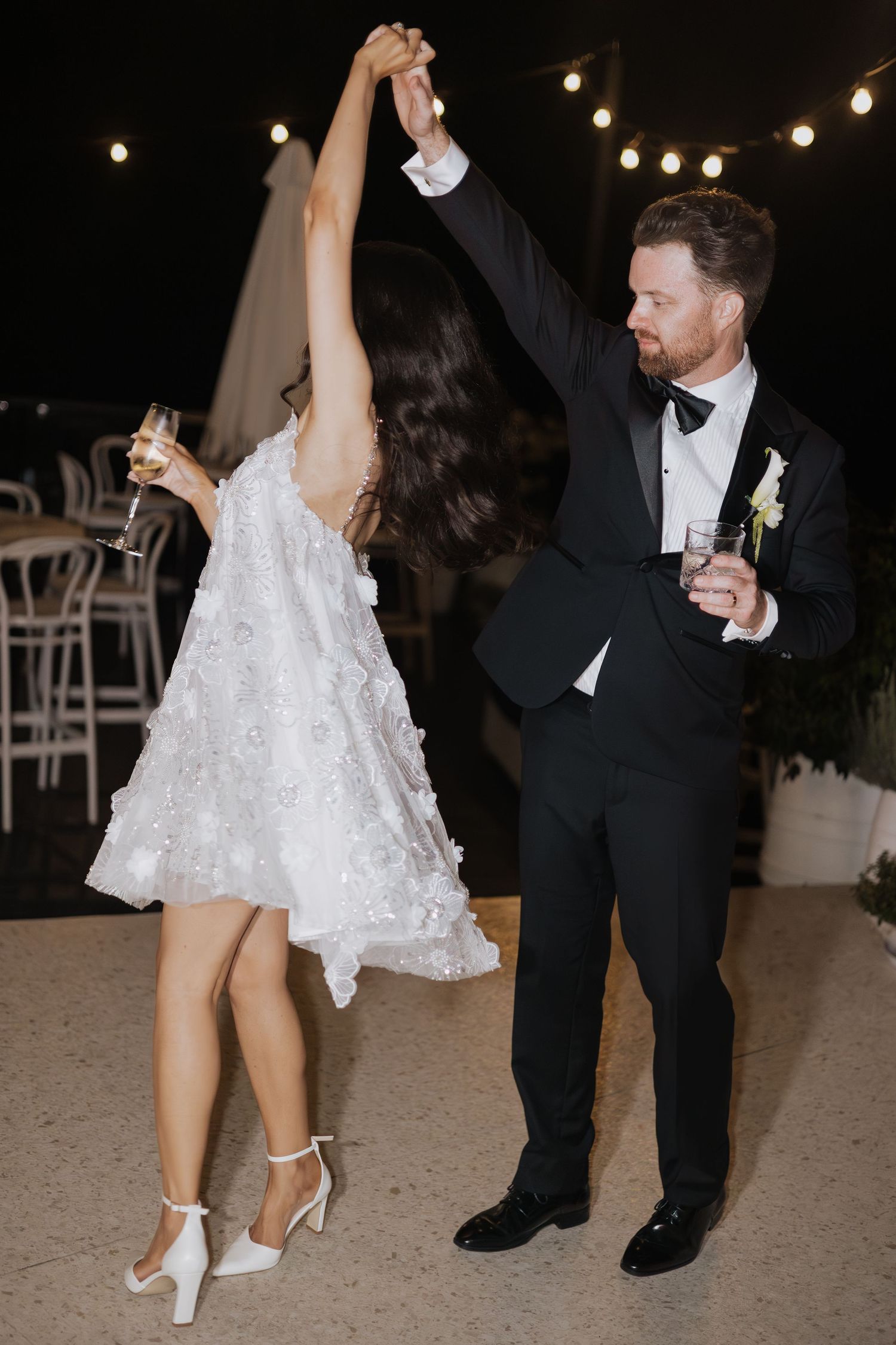 Bride and groom share first dance at outdoor wedding reception, bride in lace mini dress, groom in black tuxedo.