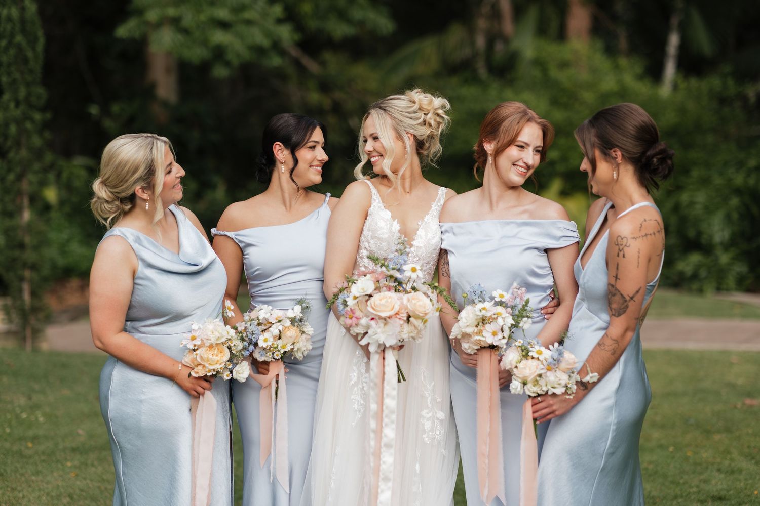 Bride in lace gown with four bridesmaids in light blue dresses holding pastel bouquets outdoors.