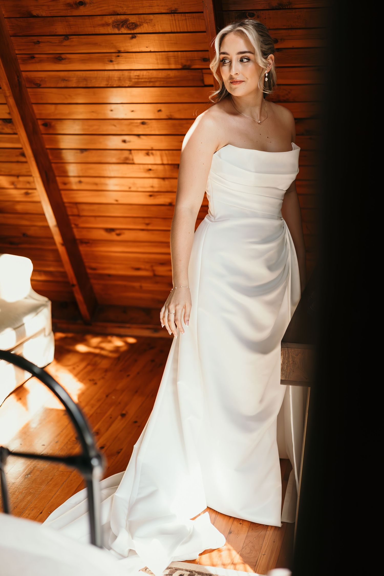 Bride in strapless white satin wedding gown poses in rustic wooden cabin room with warm sunlight.