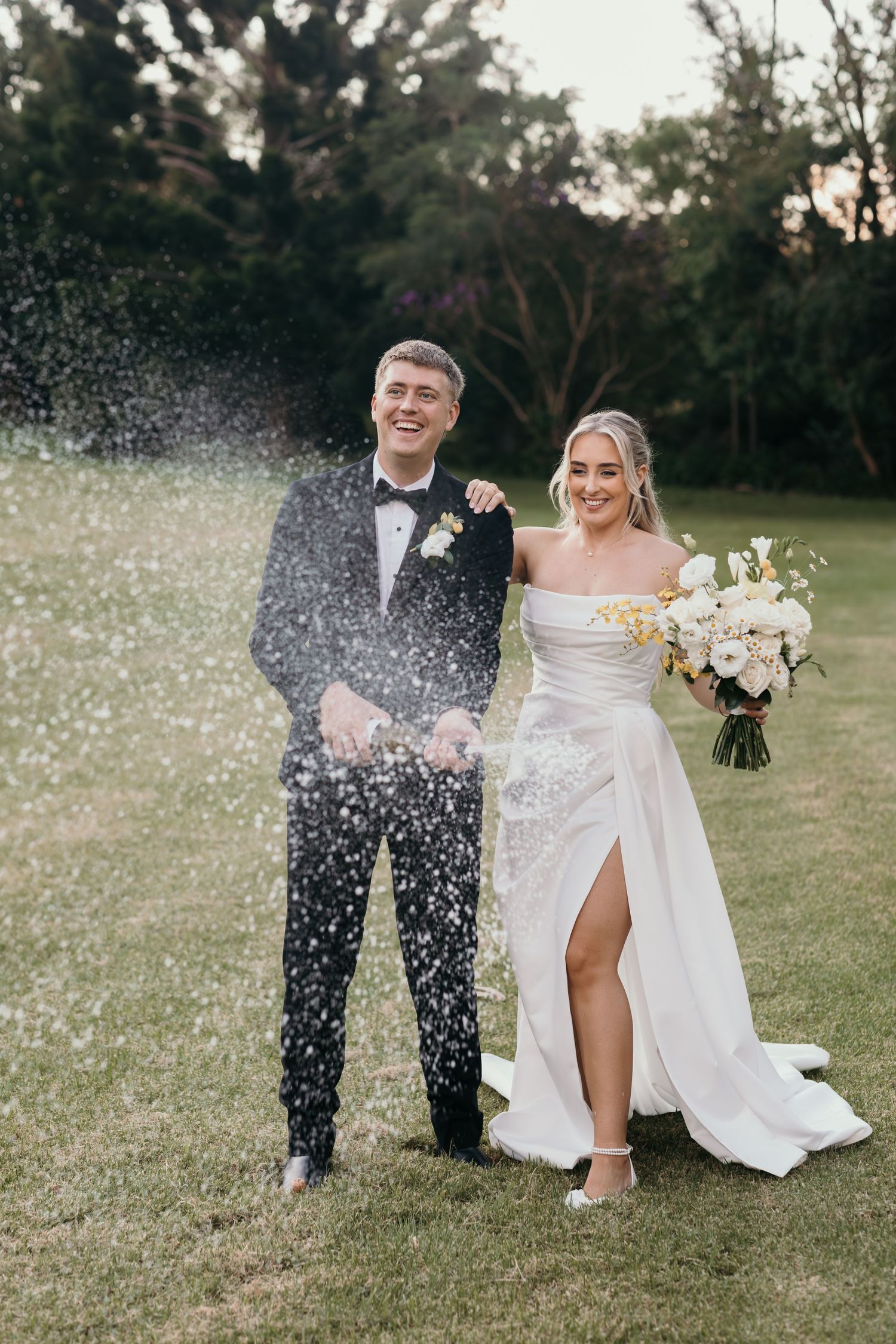 Bride and groom laughing outdoors as champagne sprays, she holds white bouquet in elegant strapless gown, he wears black tuxedo.