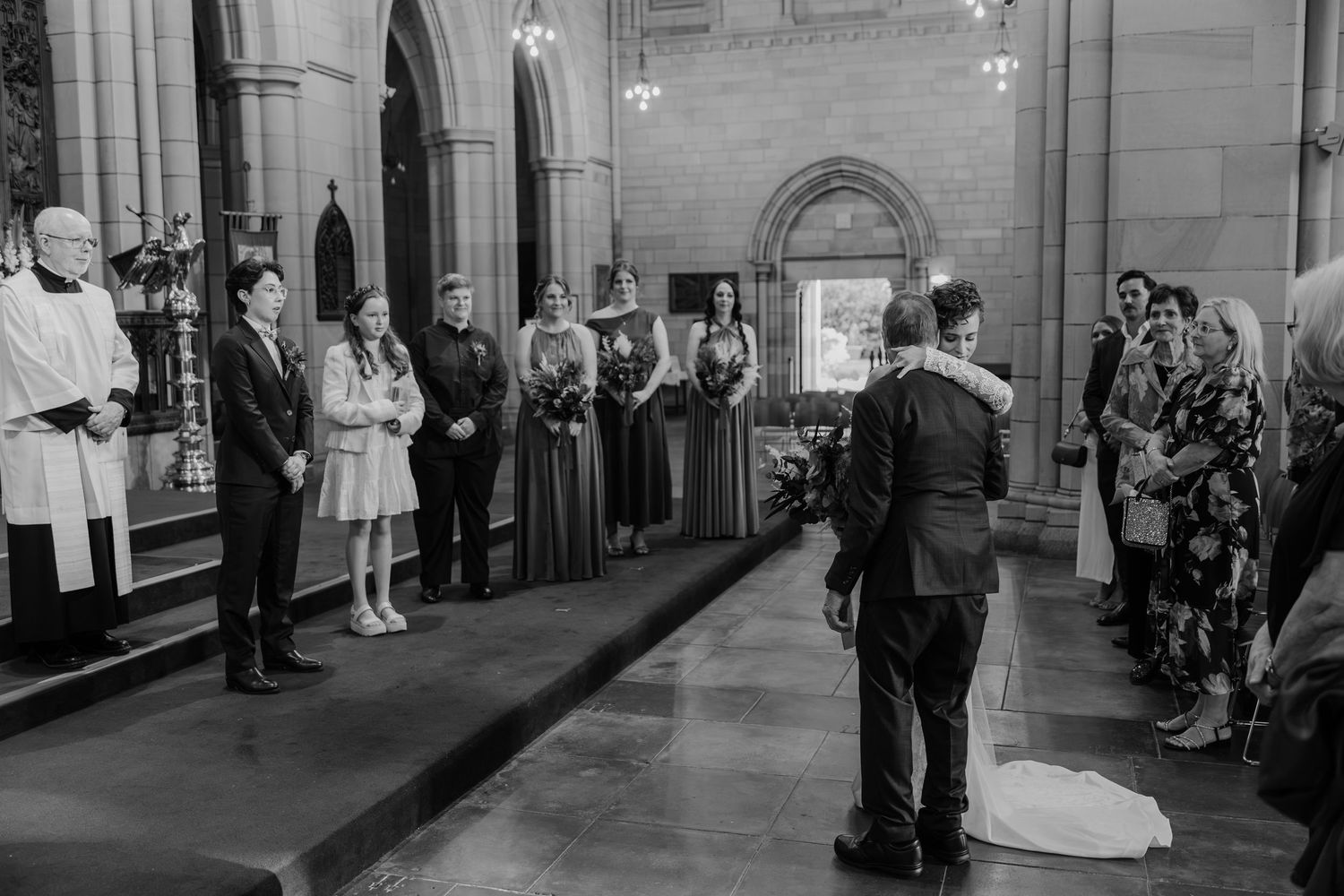 Black and white photo of a wedding ceremony inside a grand church with bridesmaids, guests, and the couple at the altar.