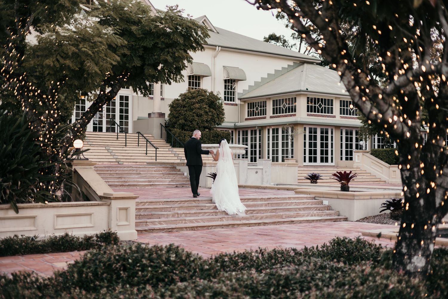 Bride and groom walk hand in hand on elegant stone steps outside a grand wedding venue adorned with twinkling lights.