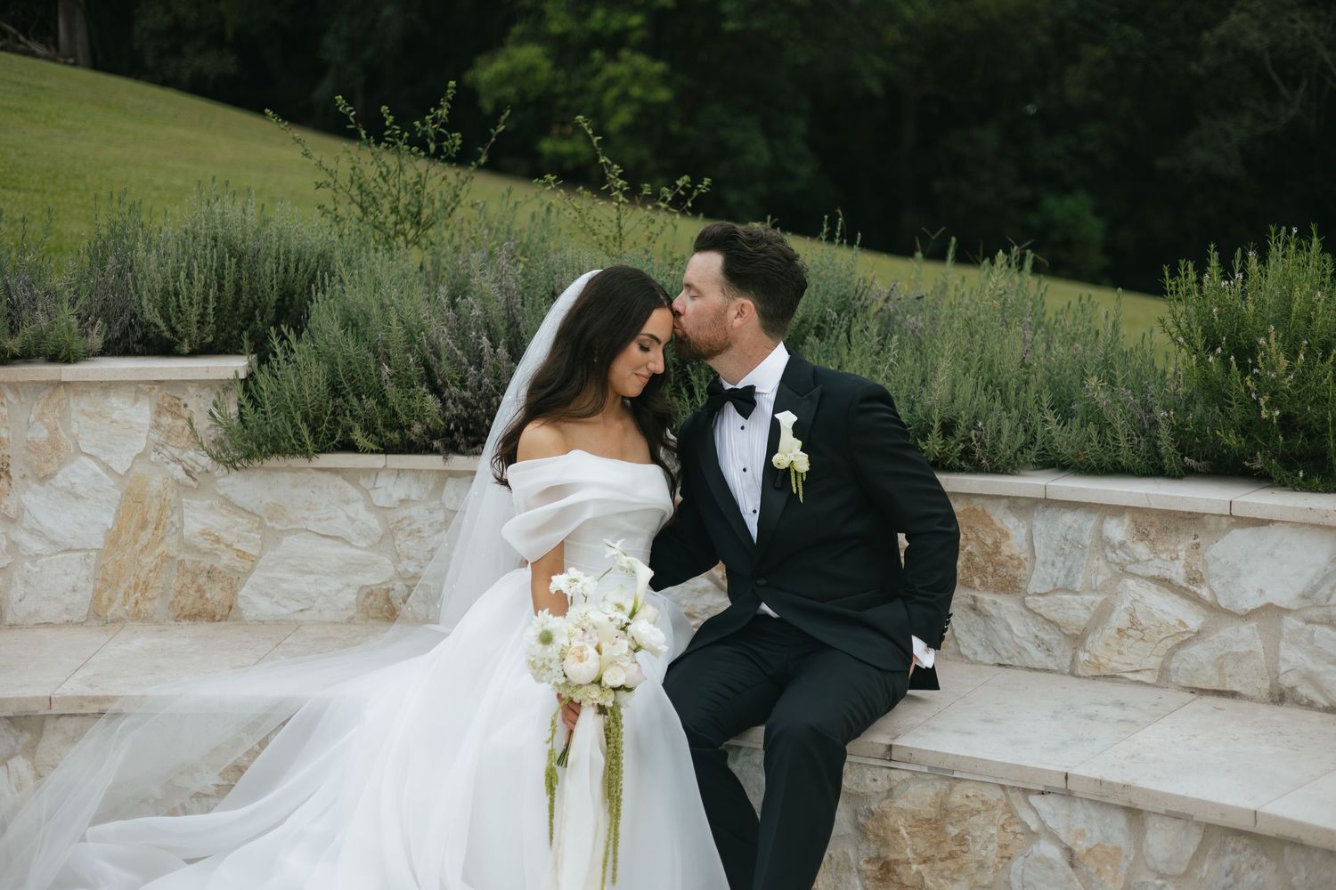 A bride and groom share a romantic kiss outdoors, she in a white off-shoulder gown with veil, he in a black tuxedo.