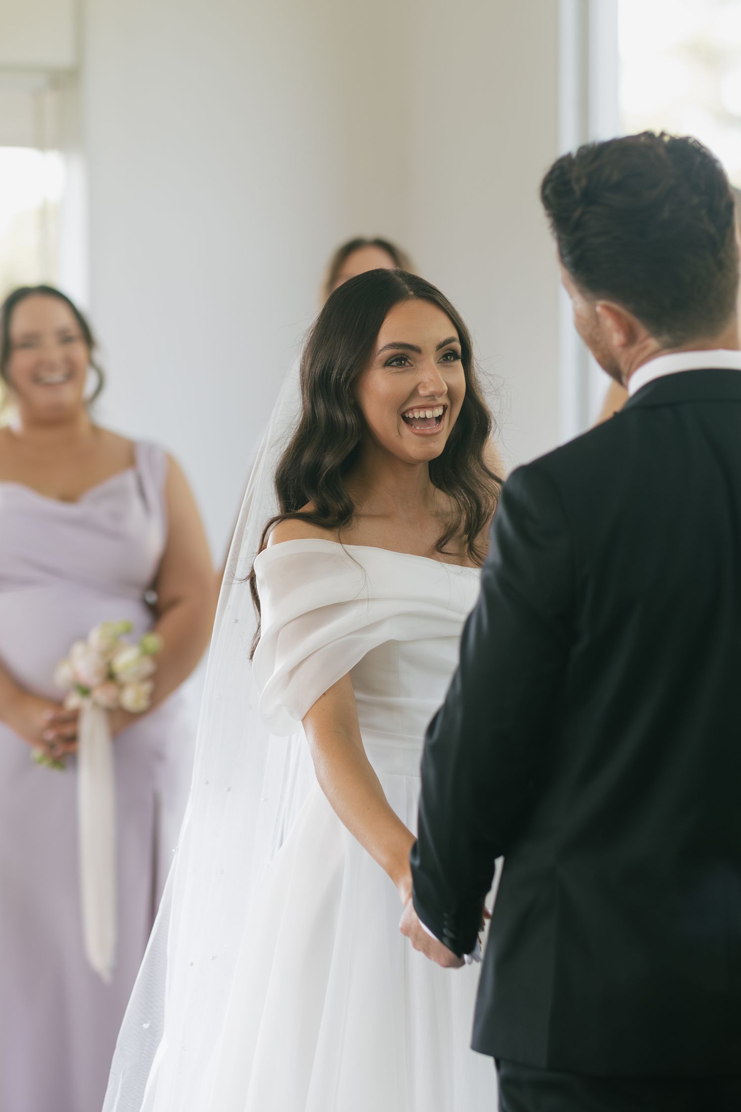 Bride in off-shoulder white gown laughing joyfully while holding hands with groom during wedding ceremony.