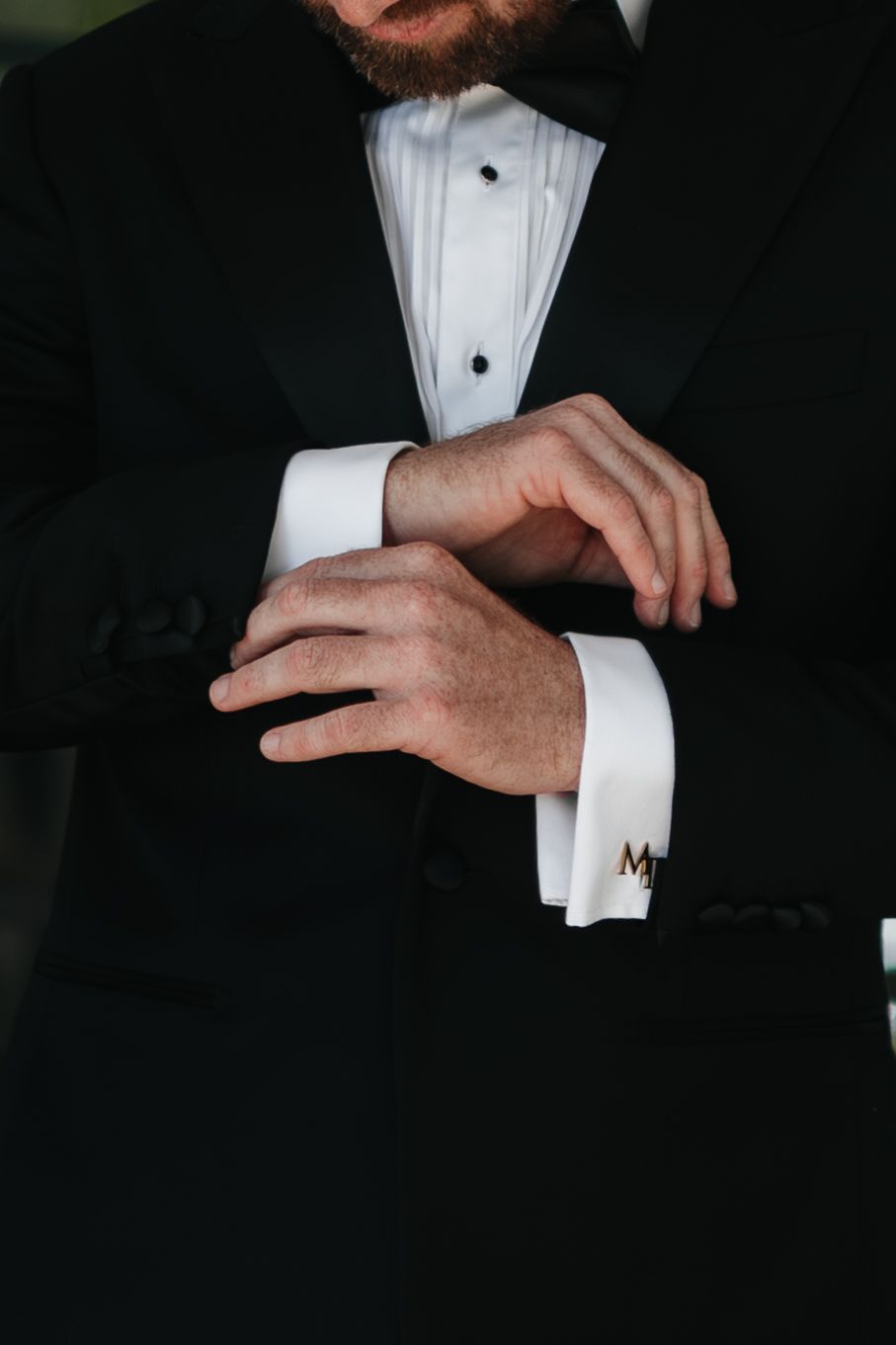 A groom in a black tuxedo adjusts his white French cuff shirt sleeve with monogrammed cufflinks.