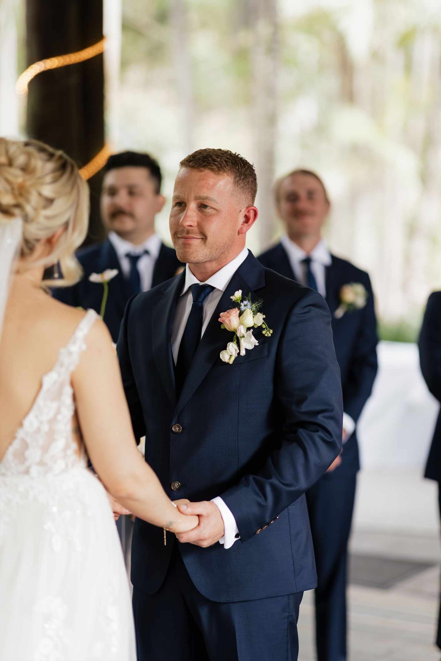 Groom in navy suit holding bride's hands at wedding ceremony with groomsmen standing in background.