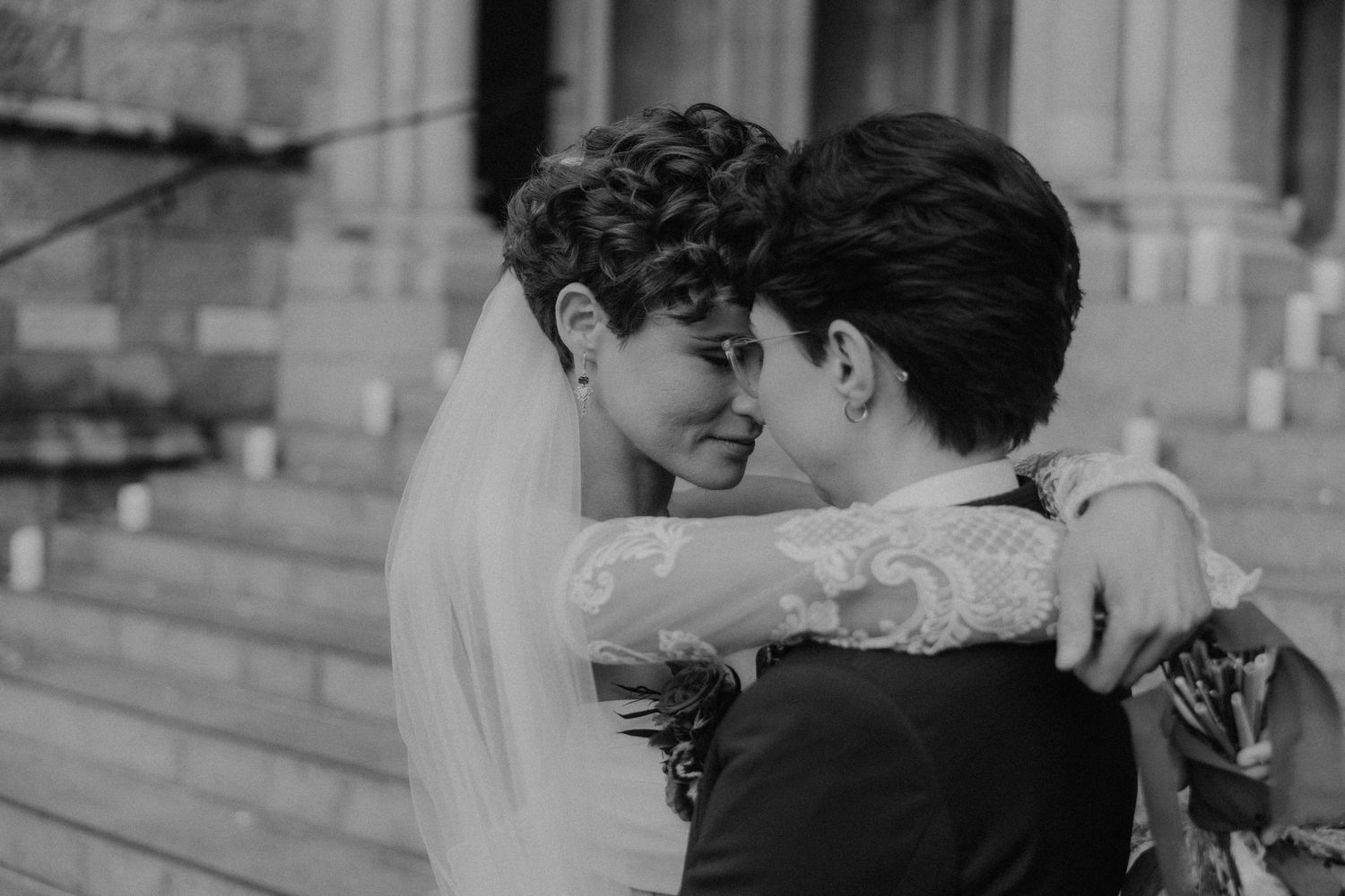 Black and white photo of two brides embracing tenderly on church steps in wedding attire with veil.