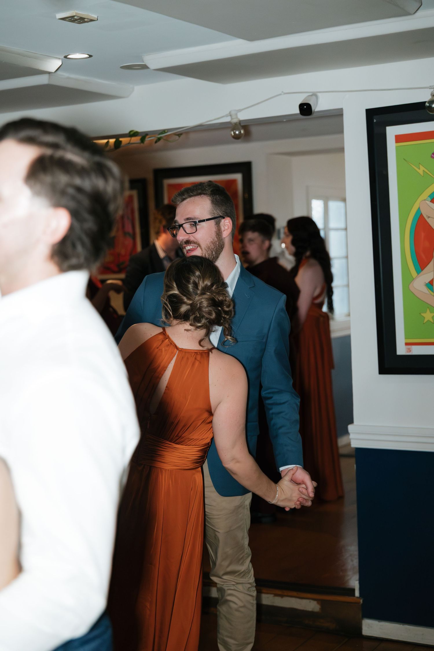 Couple holding hands at a wedding reception in a gallery space, woman in rust orange gown, man in blue blazer.