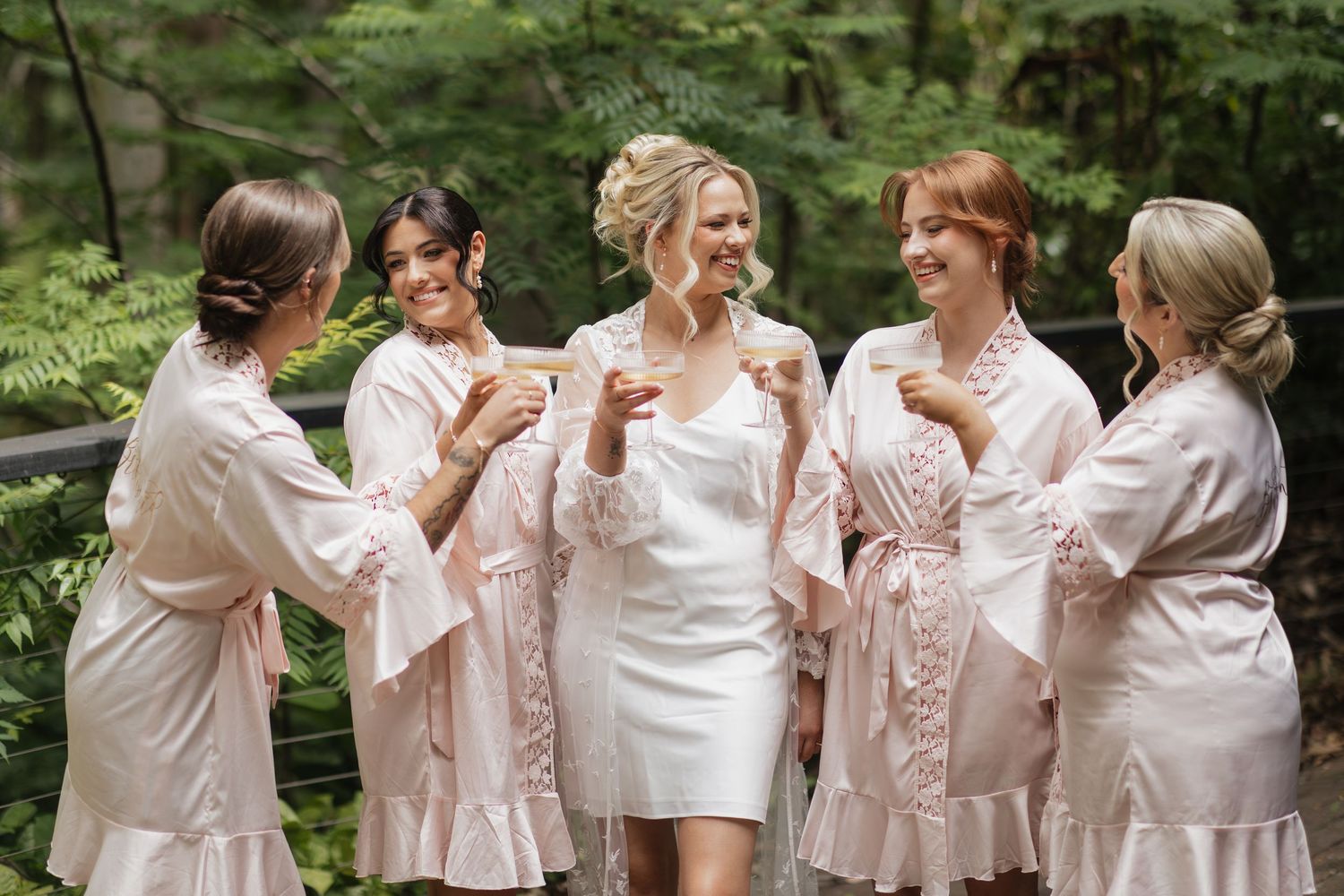 Bride in white dress laughing with four bridesmaids in pink robes outdoors among lush green foliage.
