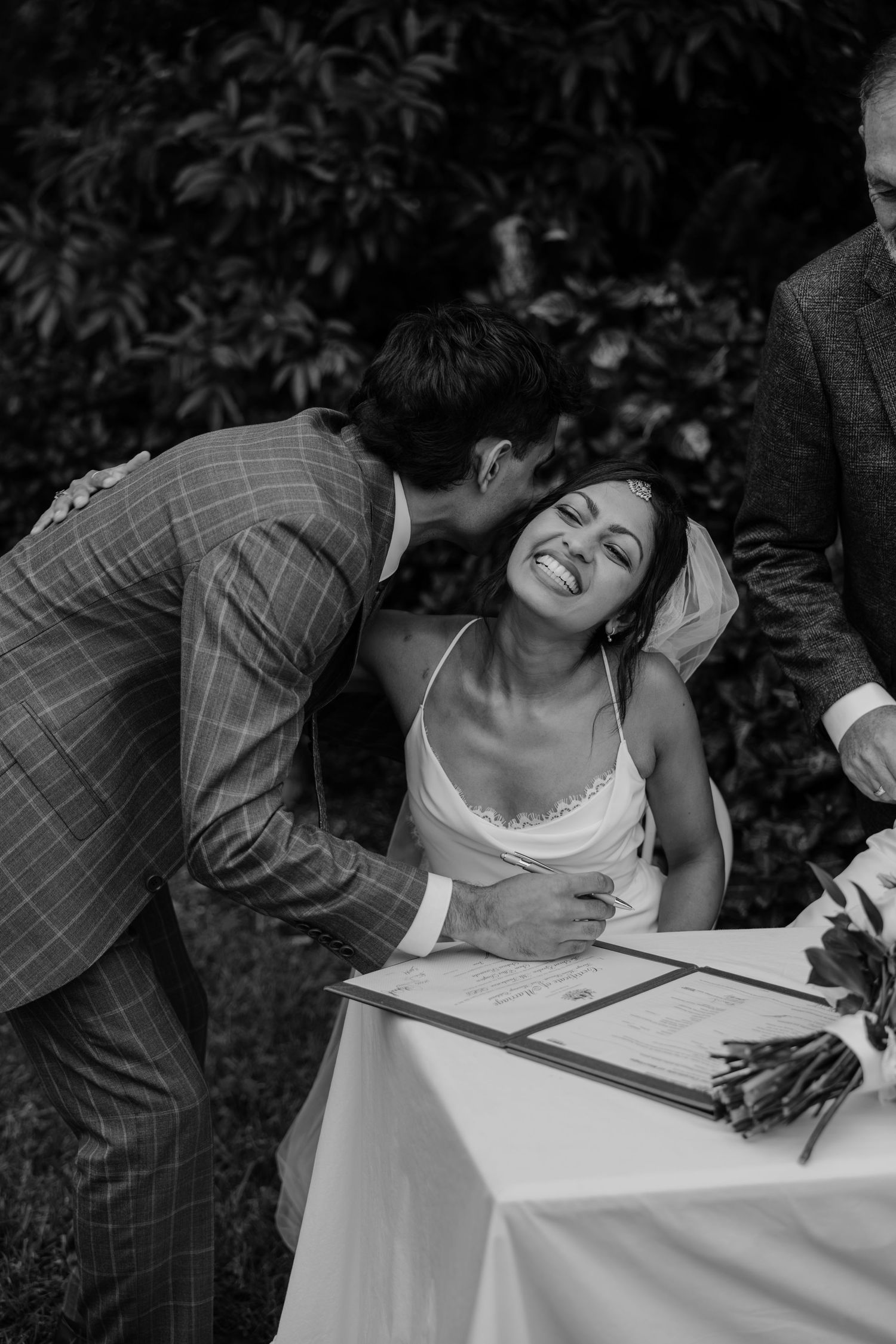 Groom in plaid suit kisses bride while she signs wedding certificate at outdoor ceremony table.