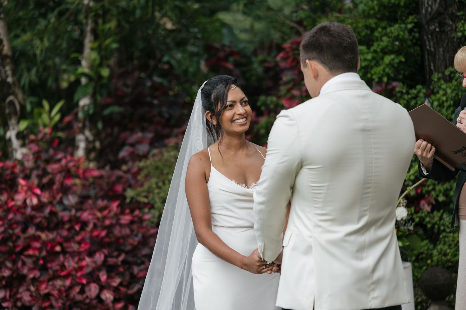 Bride in white gown and veil smiles at groom during outdoor wedding ceremony surrounded by lush greenery.
