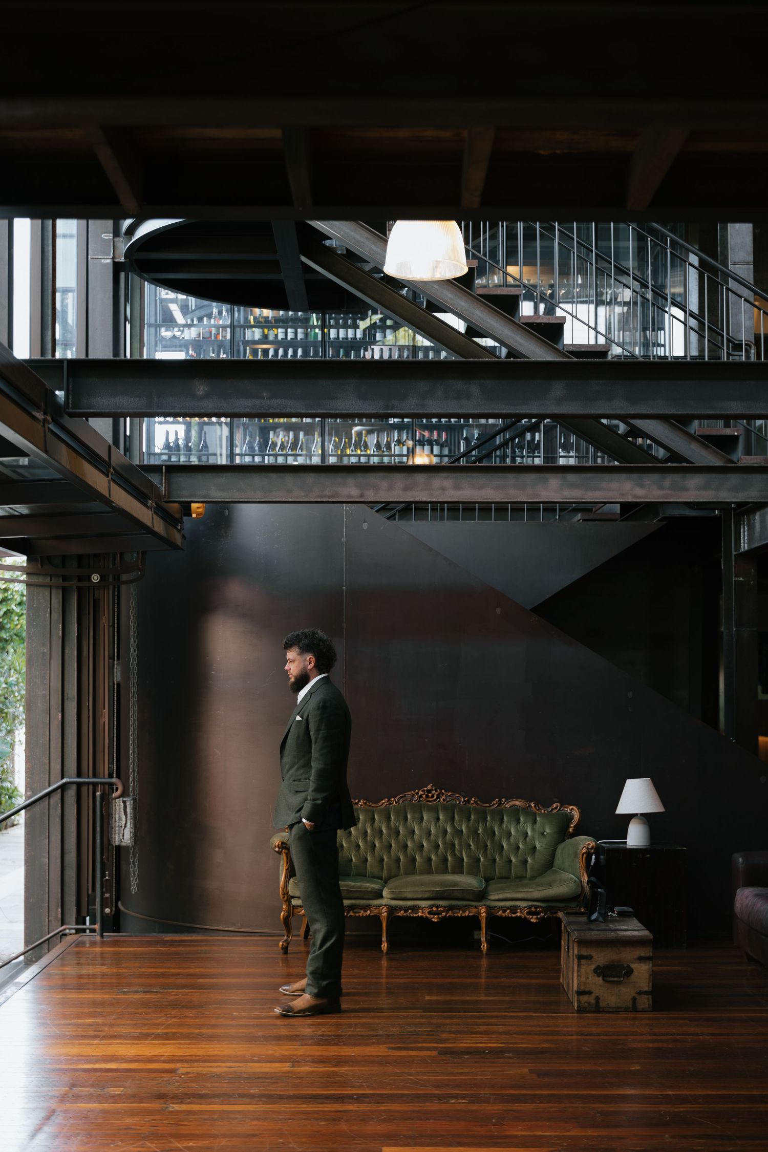 A well-dressed man in a dark suit stands in a modern industrial loft with hardwood floors and a vintage green velvet sofa.