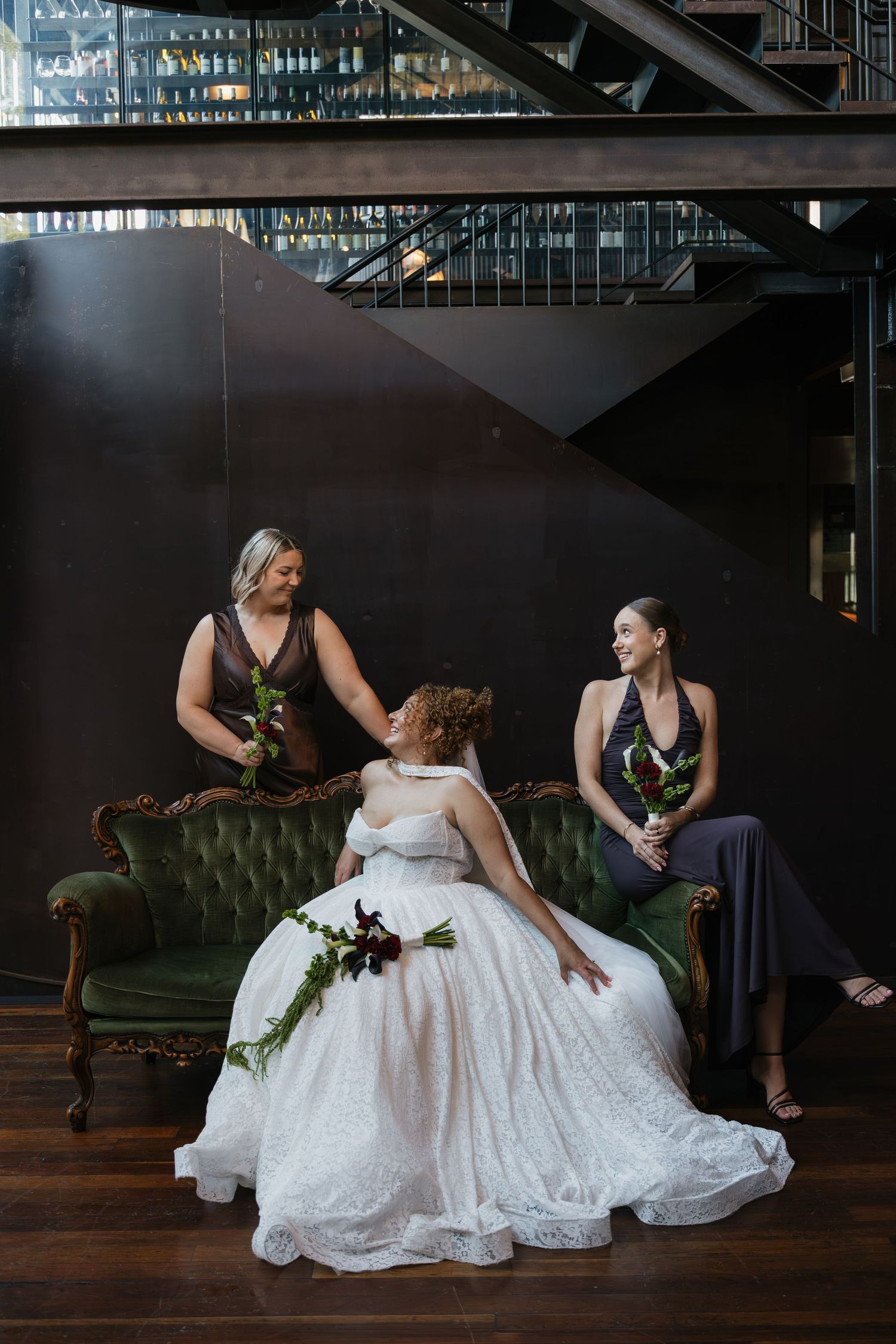 Bride in white gown sits on green velvet sofa with two bridesmaids in dark dresses, laughing together indoors.