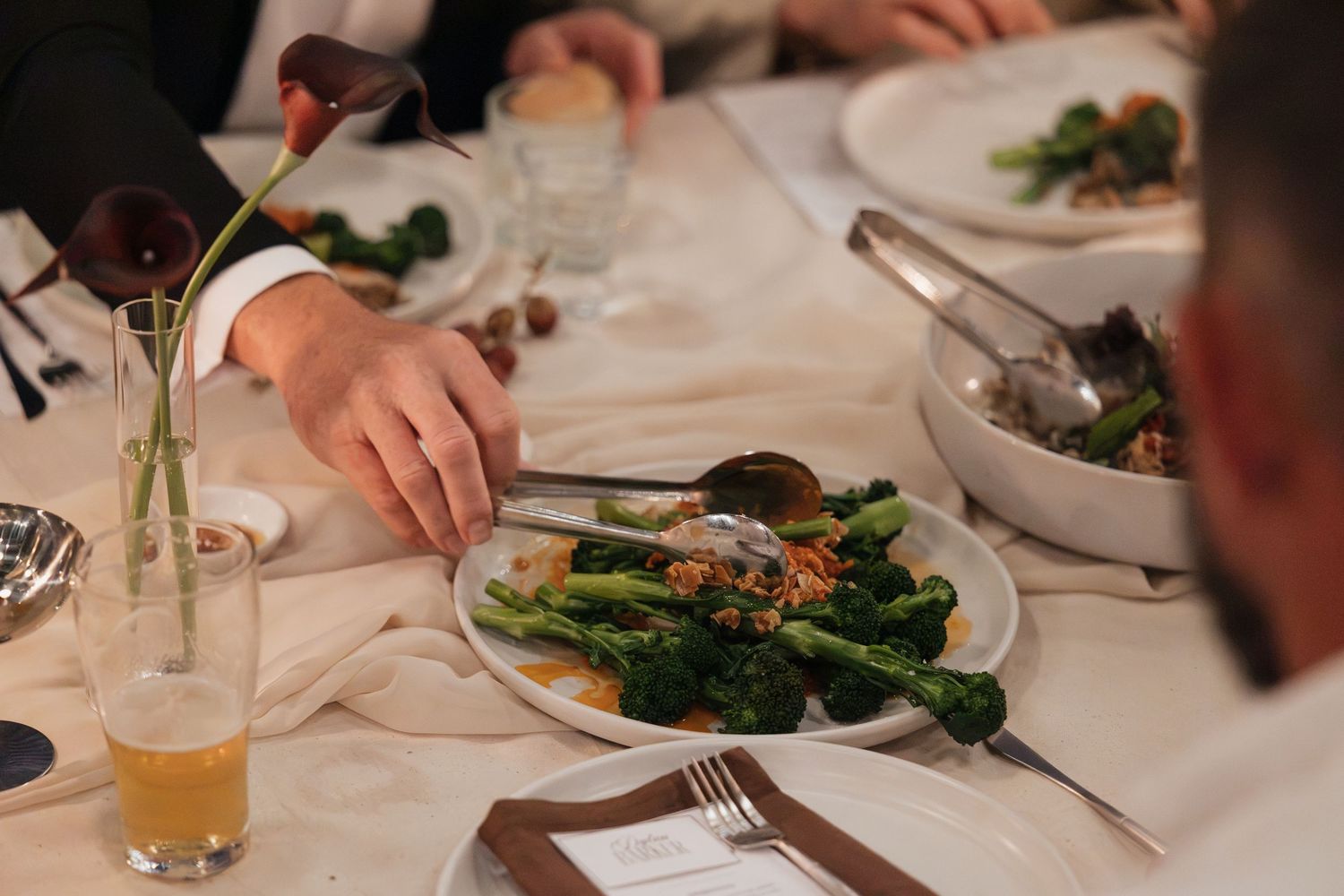 Elegant dinner table setting with broccoli dish, beer glass, and guests dining at a formal restaurant event.