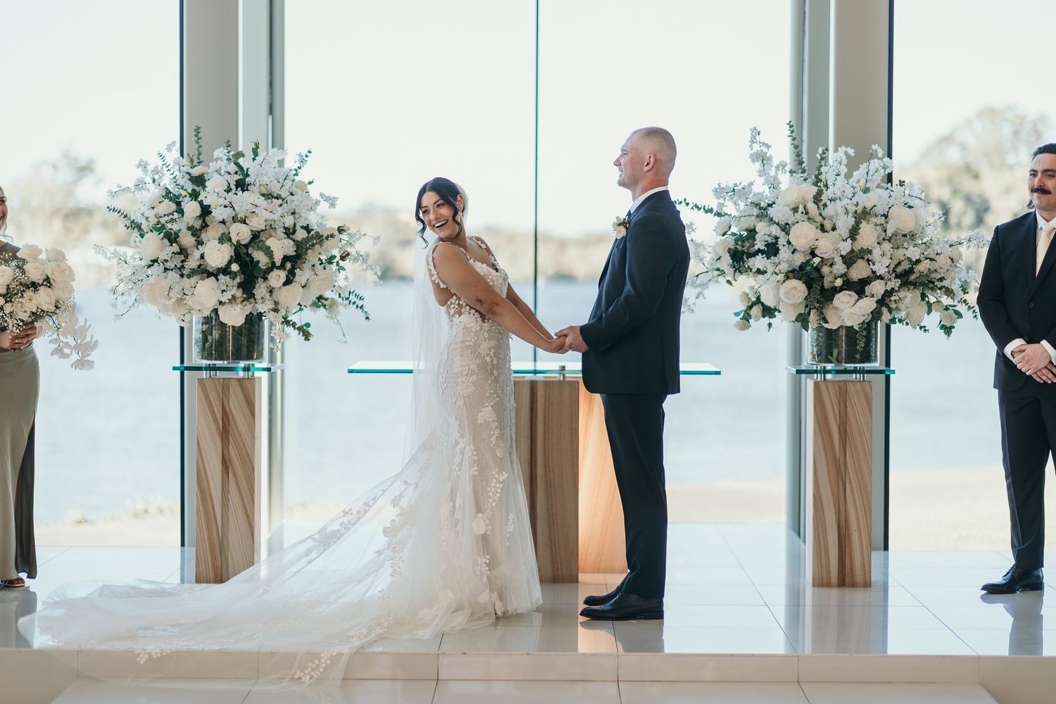 Elegant wedding ceremony setup with tall white floral arrangements flanking a modern glass-walled venue.