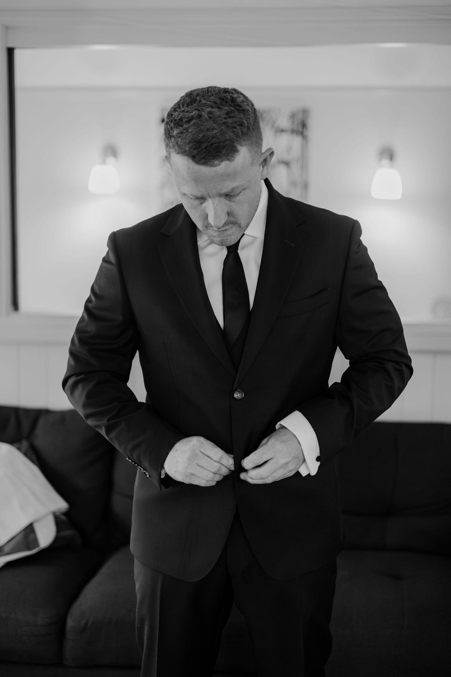 Black and white photo of a groom in a dark suit buttoning his jacket while getting ready for his wedding.
