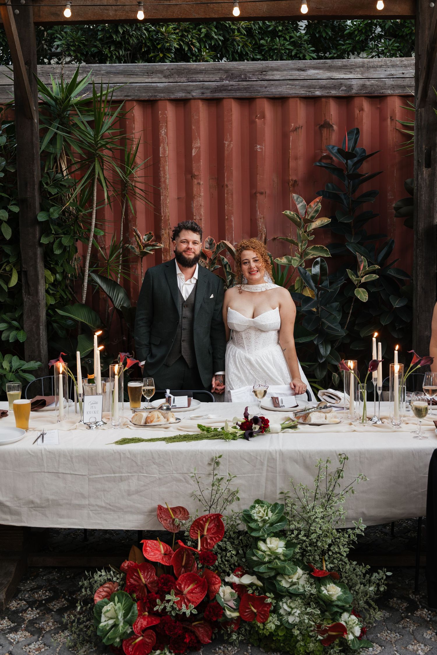 Bride and groom smiling at candlelit wedding reception table with red floral arrangements outdoors.