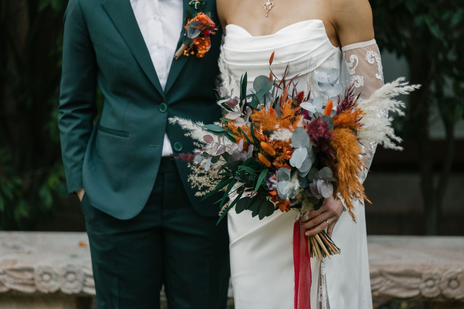 Couple in teal suit and white wedding gown holding vibrant orange and white floral bouquet with pampas grass.