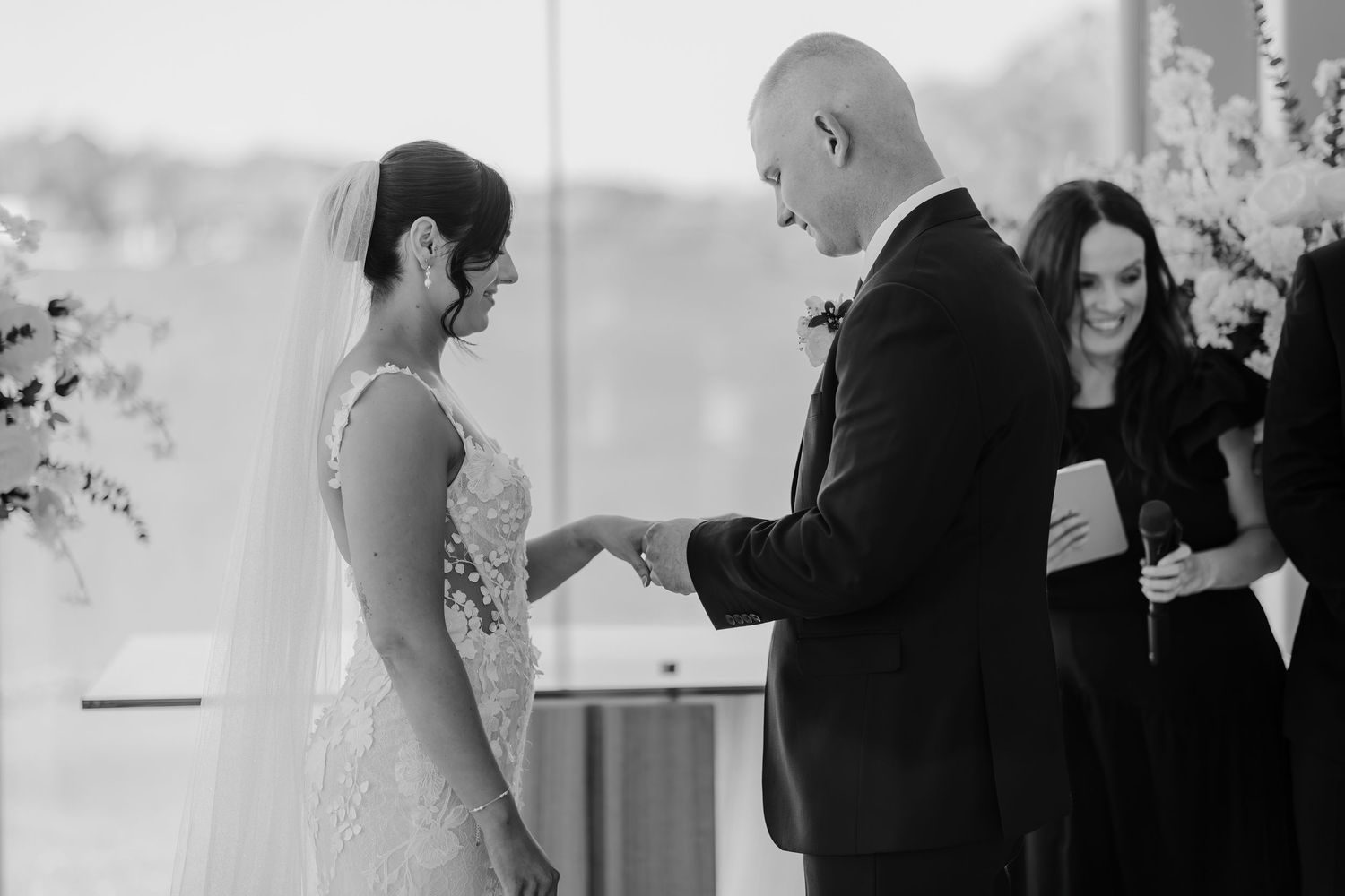Black and white photo of a wedding ceremony with a couple exchanging rings at an indoor venue.