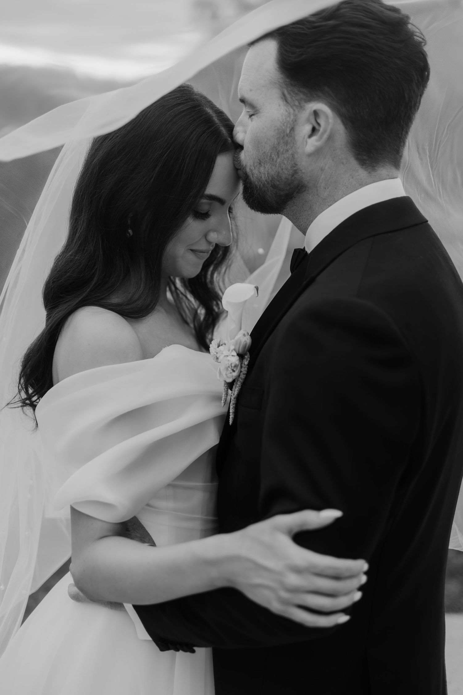 Black and white wedding photo of bride and groom sharing intimate moment, groom kissing bride's forehead under flowing veil.