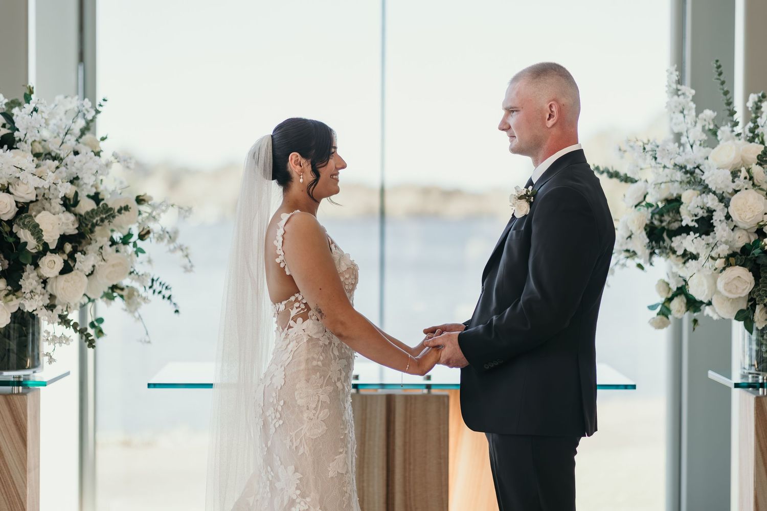 A couple exchanges vows during an elegant indoor wedding ceremony surrounded by white floral arrangements.