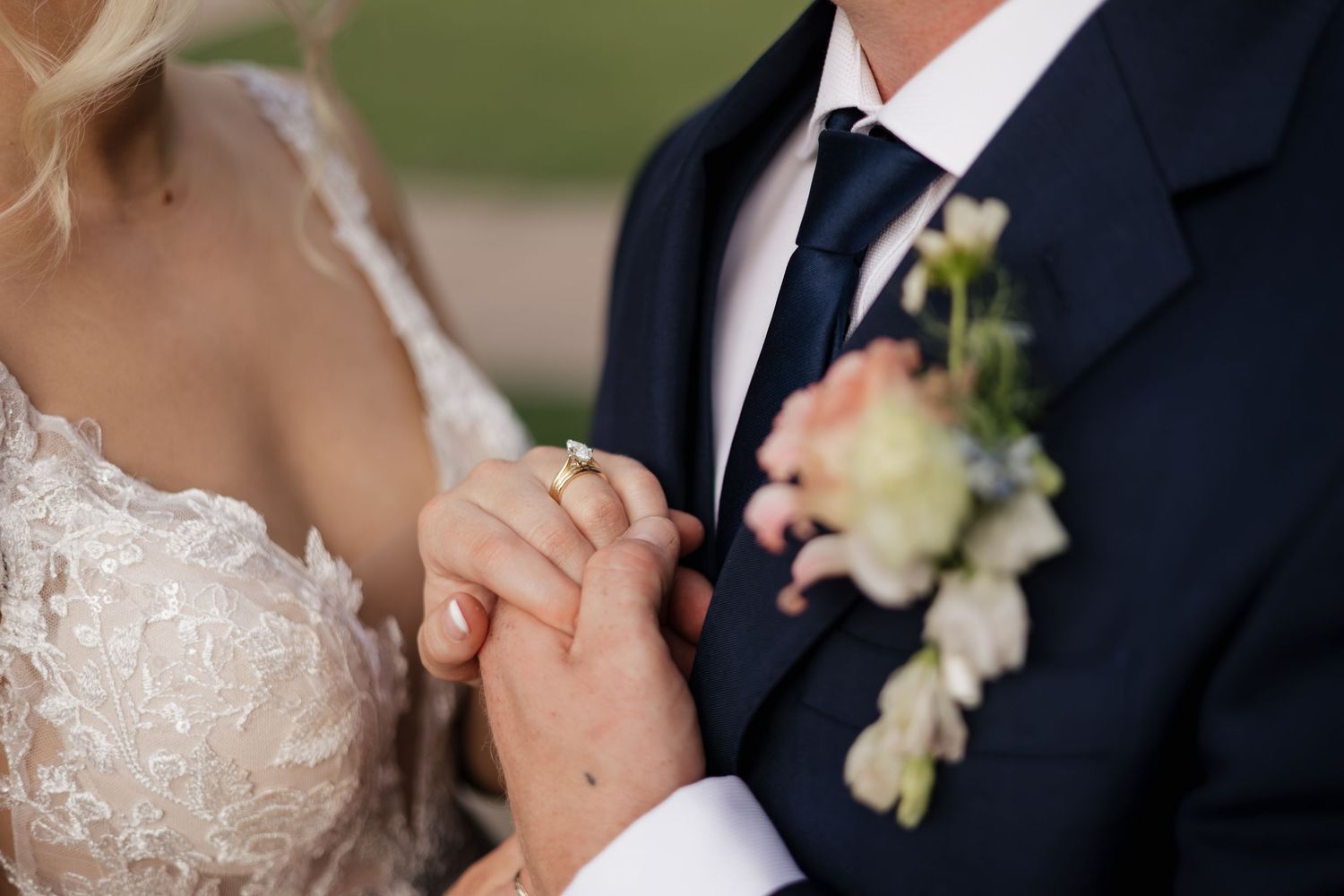Bride and groom holding hands, showcasing wedding ring, groom wearing navy suit with floral boutonniere.