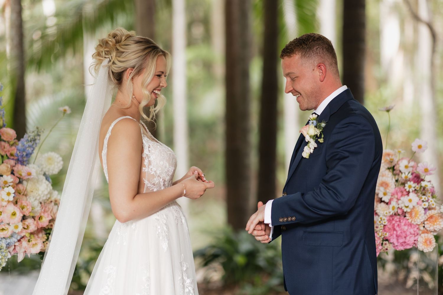 Bride and groom exchanging vows outdoors among tall trees and pastel floral arrangements at a forest wedding.