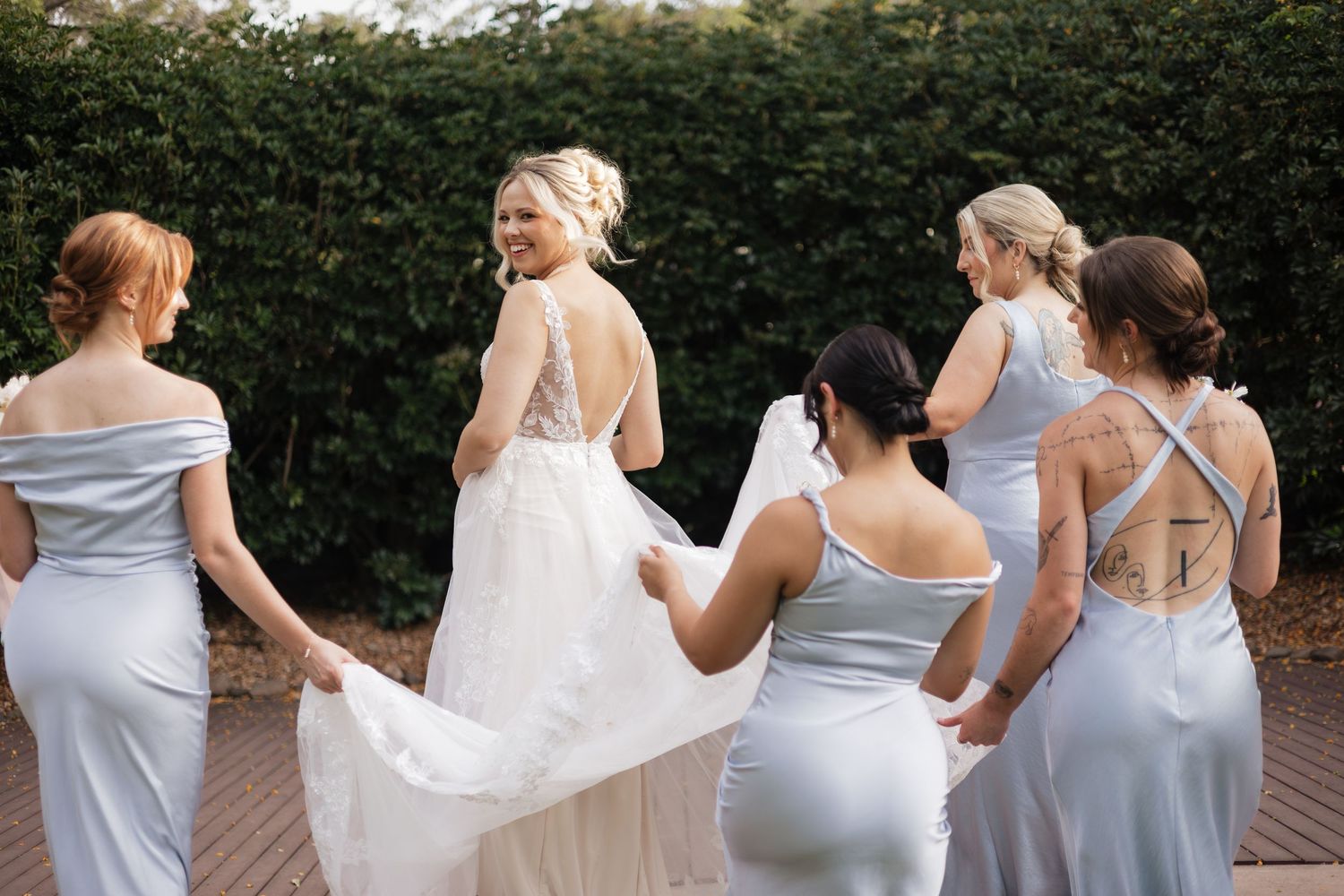 Bride in white gown laughing with bridesmaids in silver dresses holding her train outdoors.