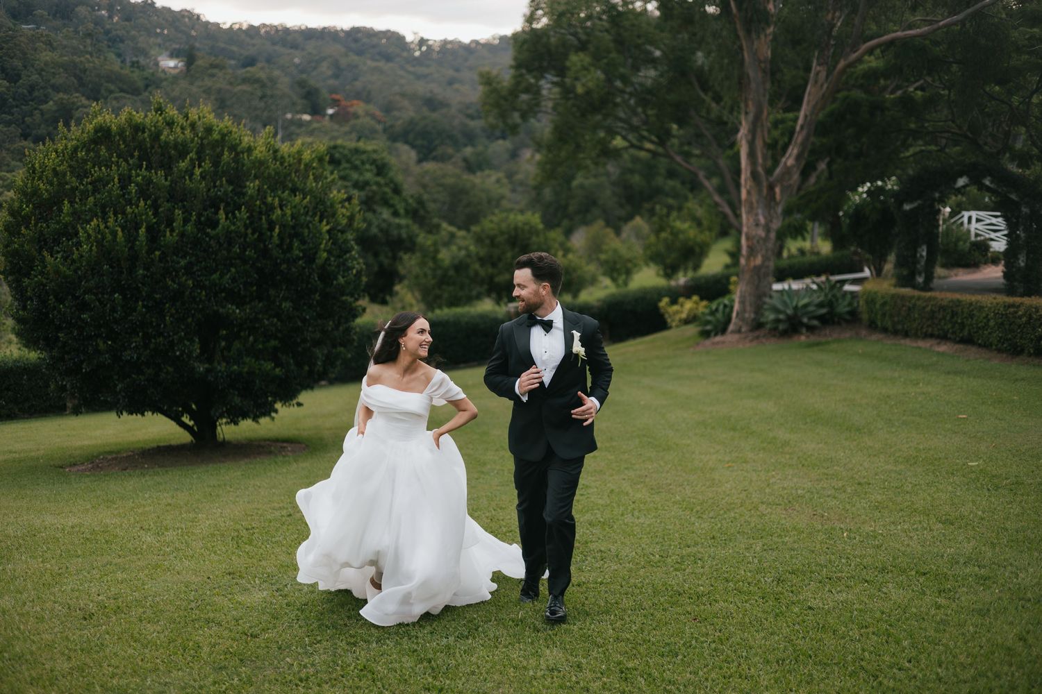 Bride and groom walking hand in hand on lush green lawn with trees and hills in background at outdoor wedding.