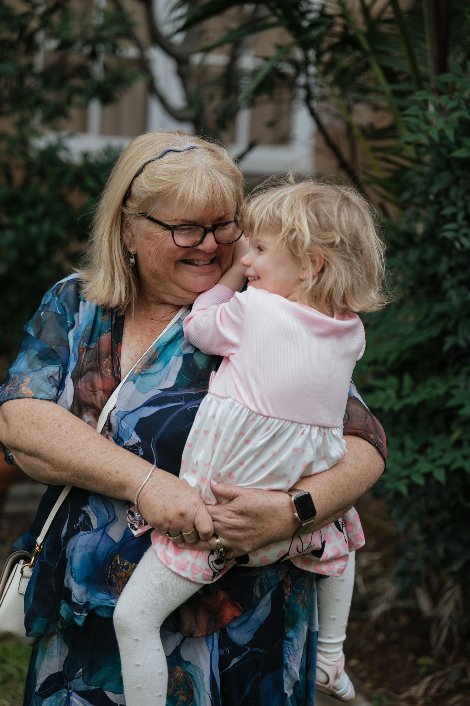 Grandmother in floral dress holding young blonde toddler girl in pink outfit, sharing a tender moment outdoors.