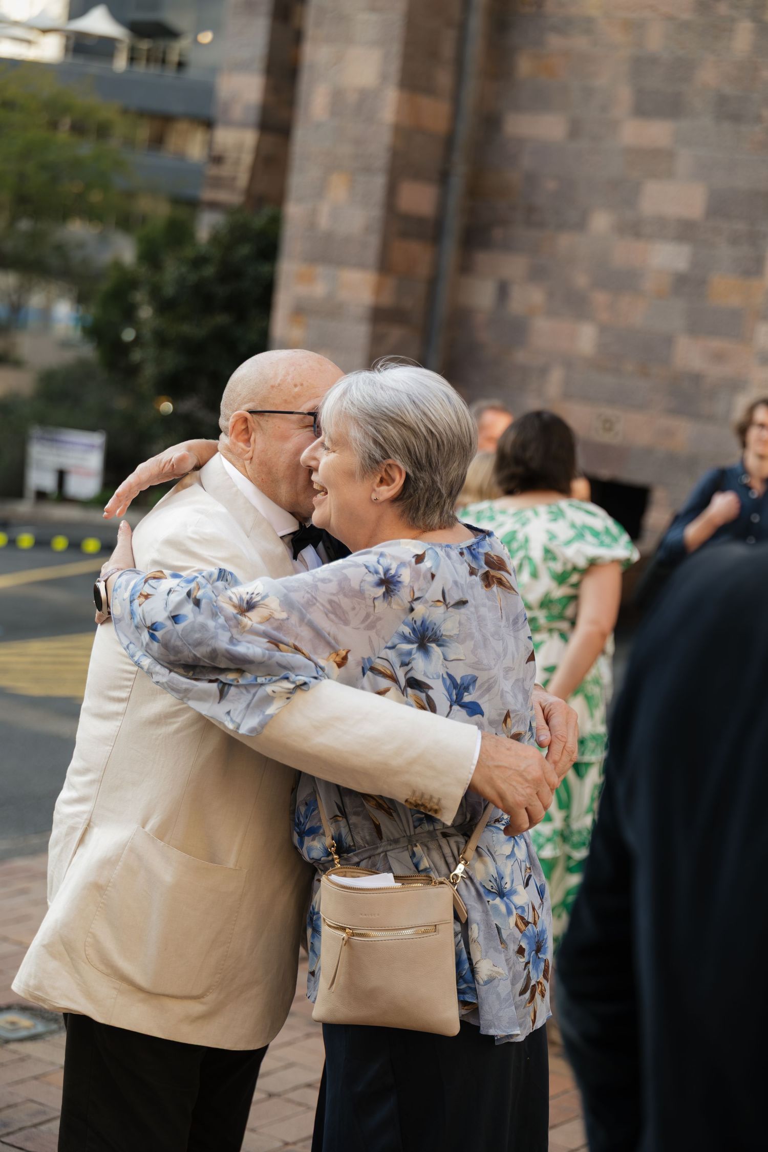 Elderly couple sharing a warm embrace outside a stone building at a formal event.