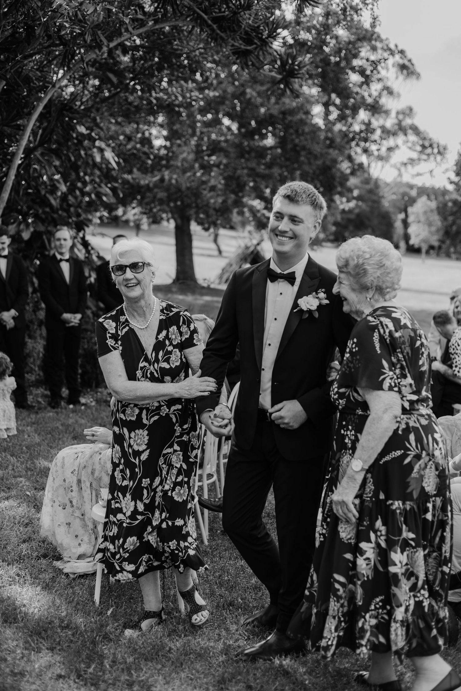 Groom in tuxedo escorts two elderly women in floral dresses at an outdoor wedding ceremony.