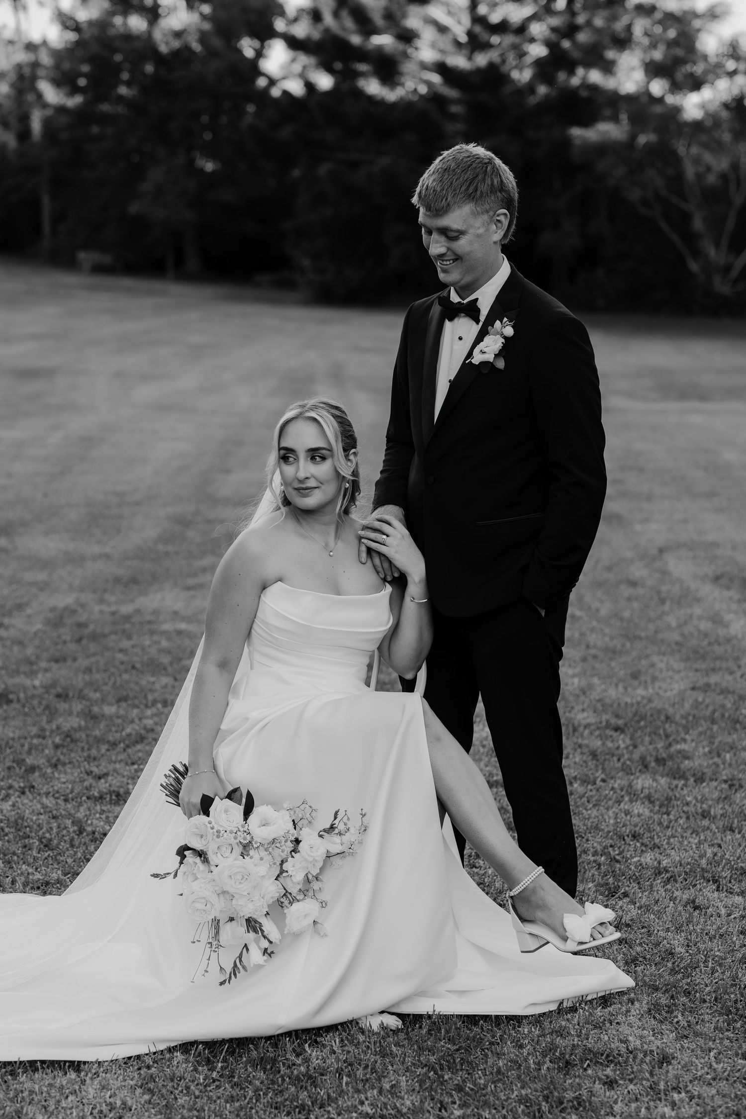 Black and white wedding photo of bride sitting on grass holding bouquet while groom in tuxedo stands beside her.