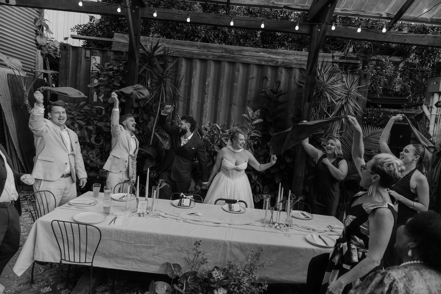Black and white photo of wedding party celebrating at outdoor reception table with string lights and tropical greenery.