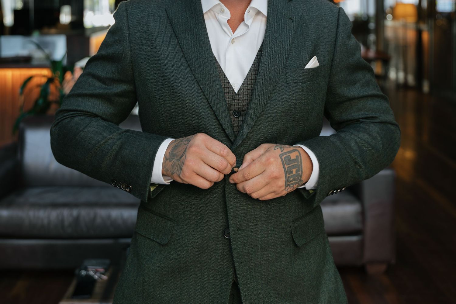 Man in green plaid suit adjusting jacket buttons, showcasing tattooed hands and white pocket square.