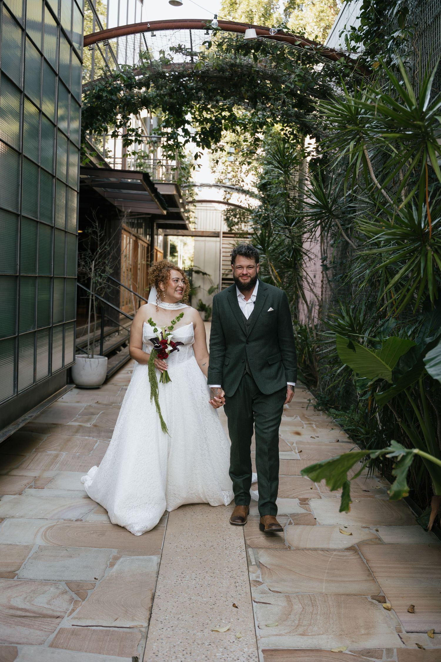 Bride and groom walk hand-in-hand through a lush garden archway, bride in white gown holding bouquet, groom in dark suit.