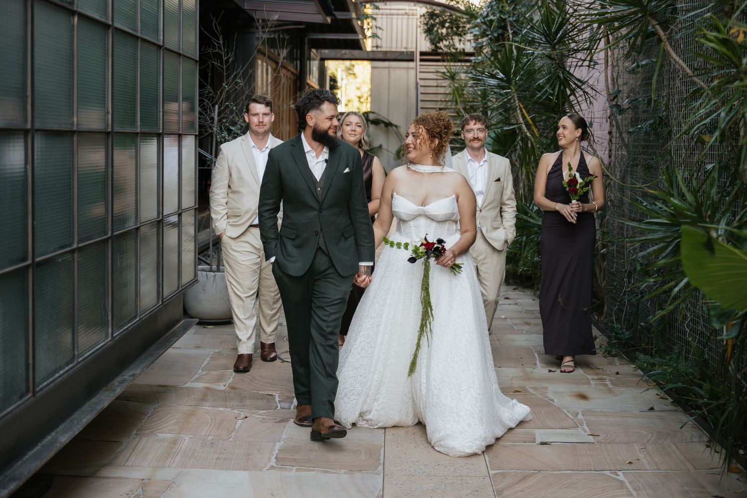 Bride and groom walk together through courtyard with wedding party following behind during outdoor ceremony.