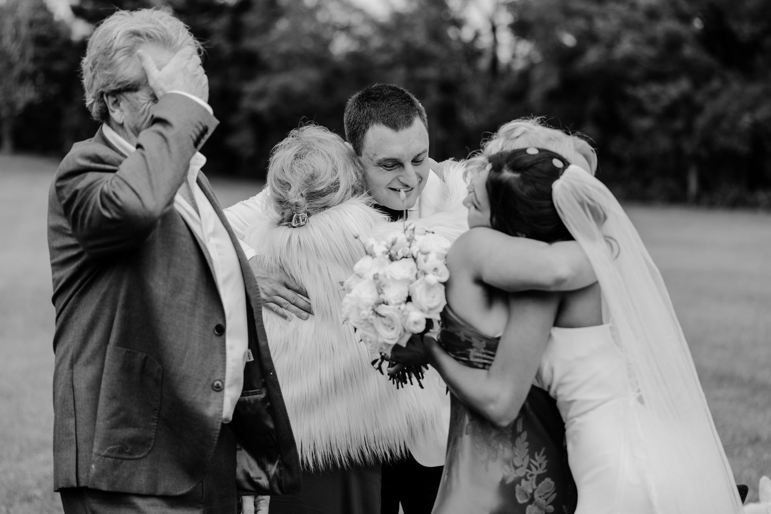 Black and white wedding photo of bride and groom sharing emotional hugs with family outdoors.