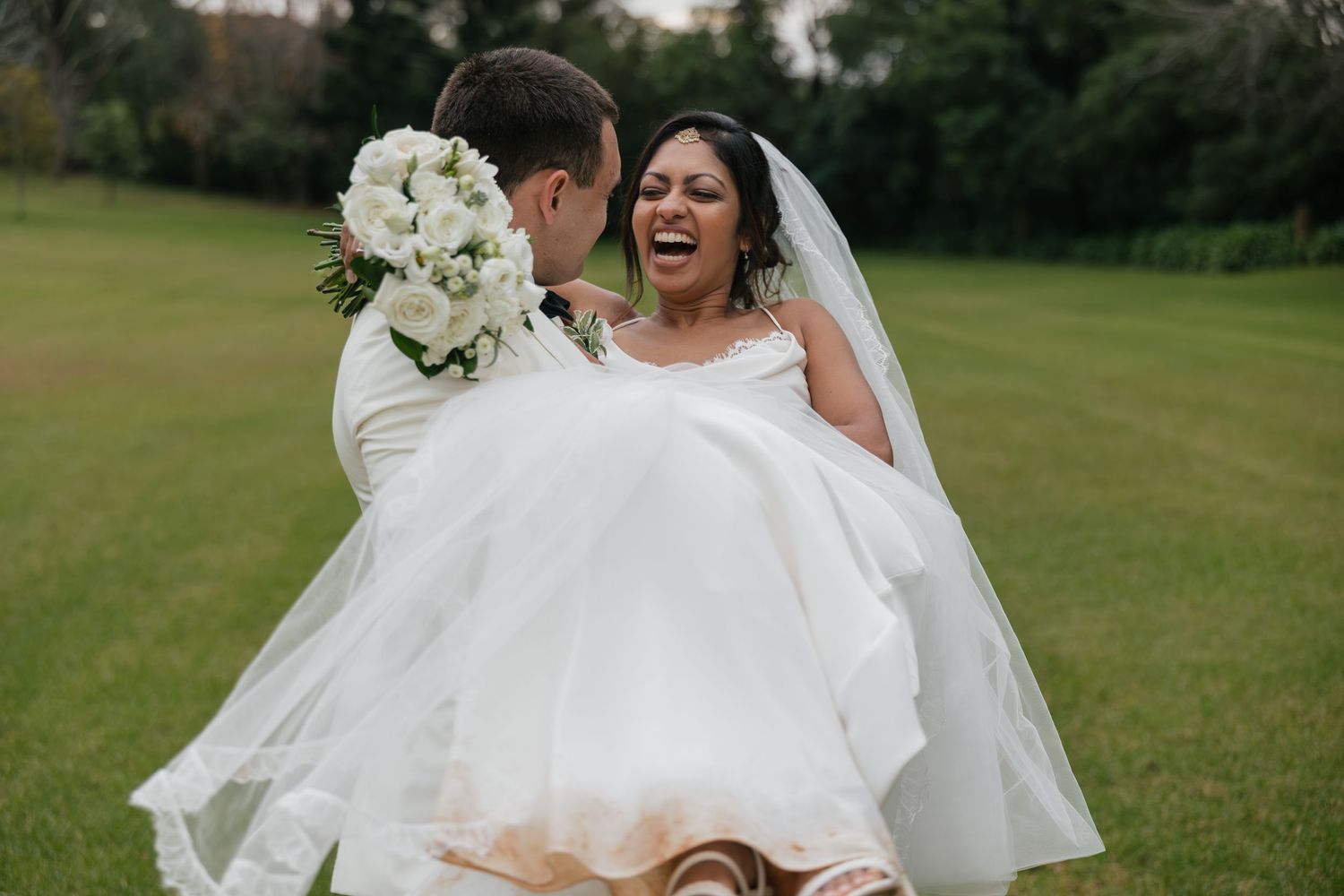 Groom lifting laughing bride in white wedding dress and veil on green lawn, holding white floral bouquet.