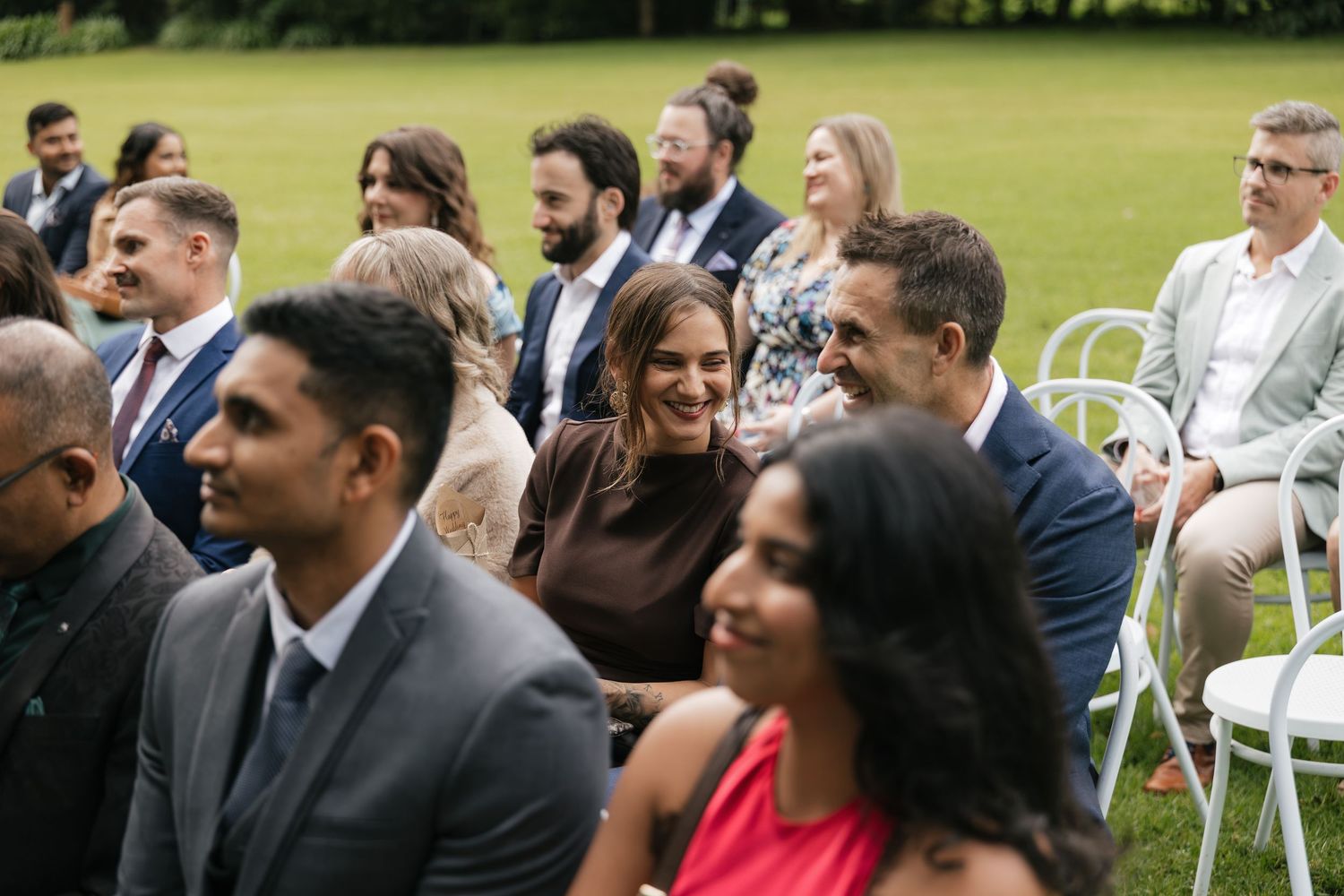 Guests smiling and laughing during an outdoor wedding ceremony seated on white chairs on a green lawn.