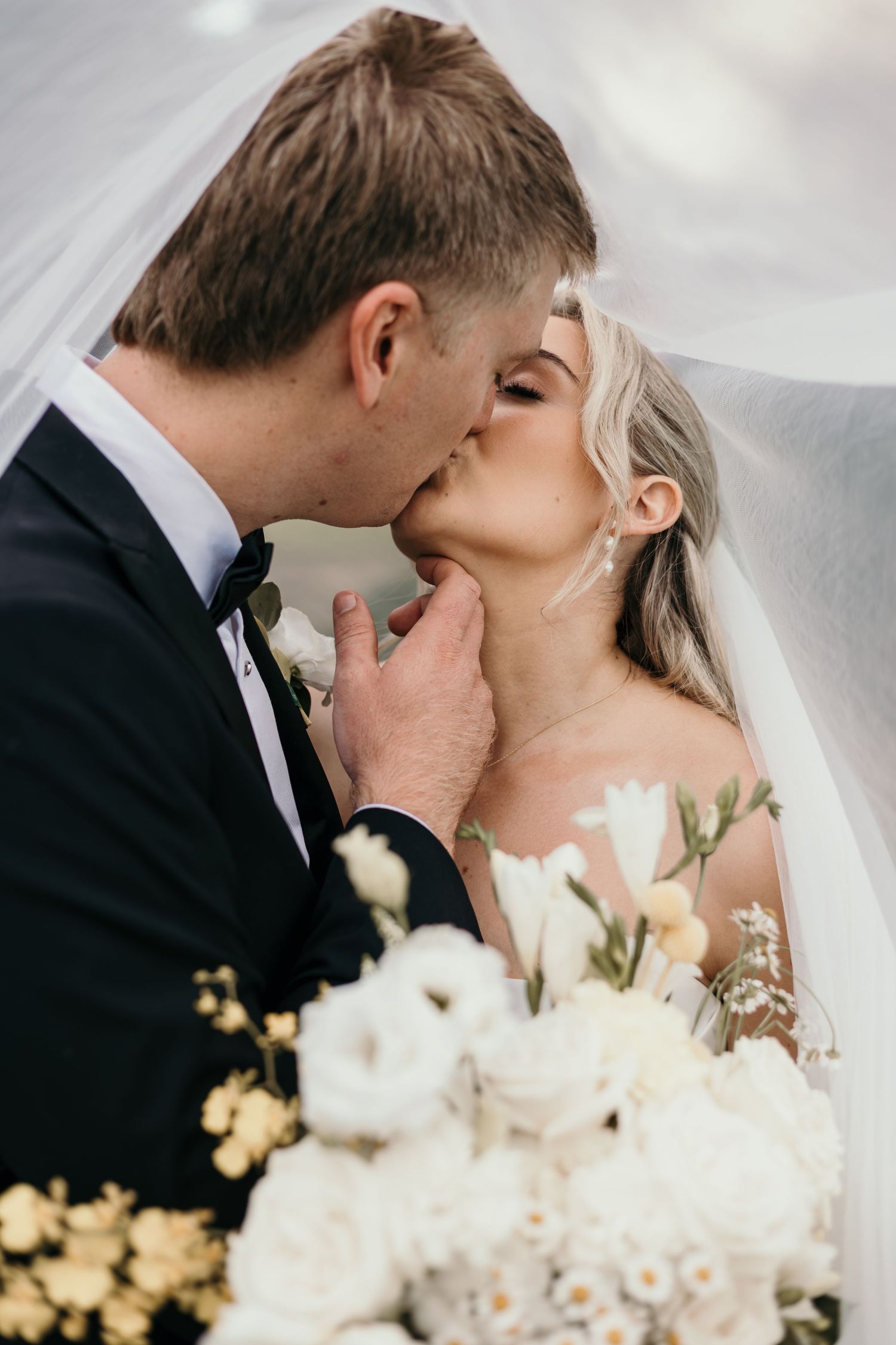 A groom in a black tuxedo tenderly kisses his bride under a veil, with a white floral bouquet in the foreground.