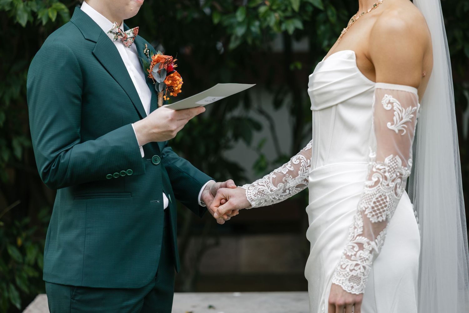 Groom in green suit reads vows to bride in lace wedding dress during outdoor ceremony.