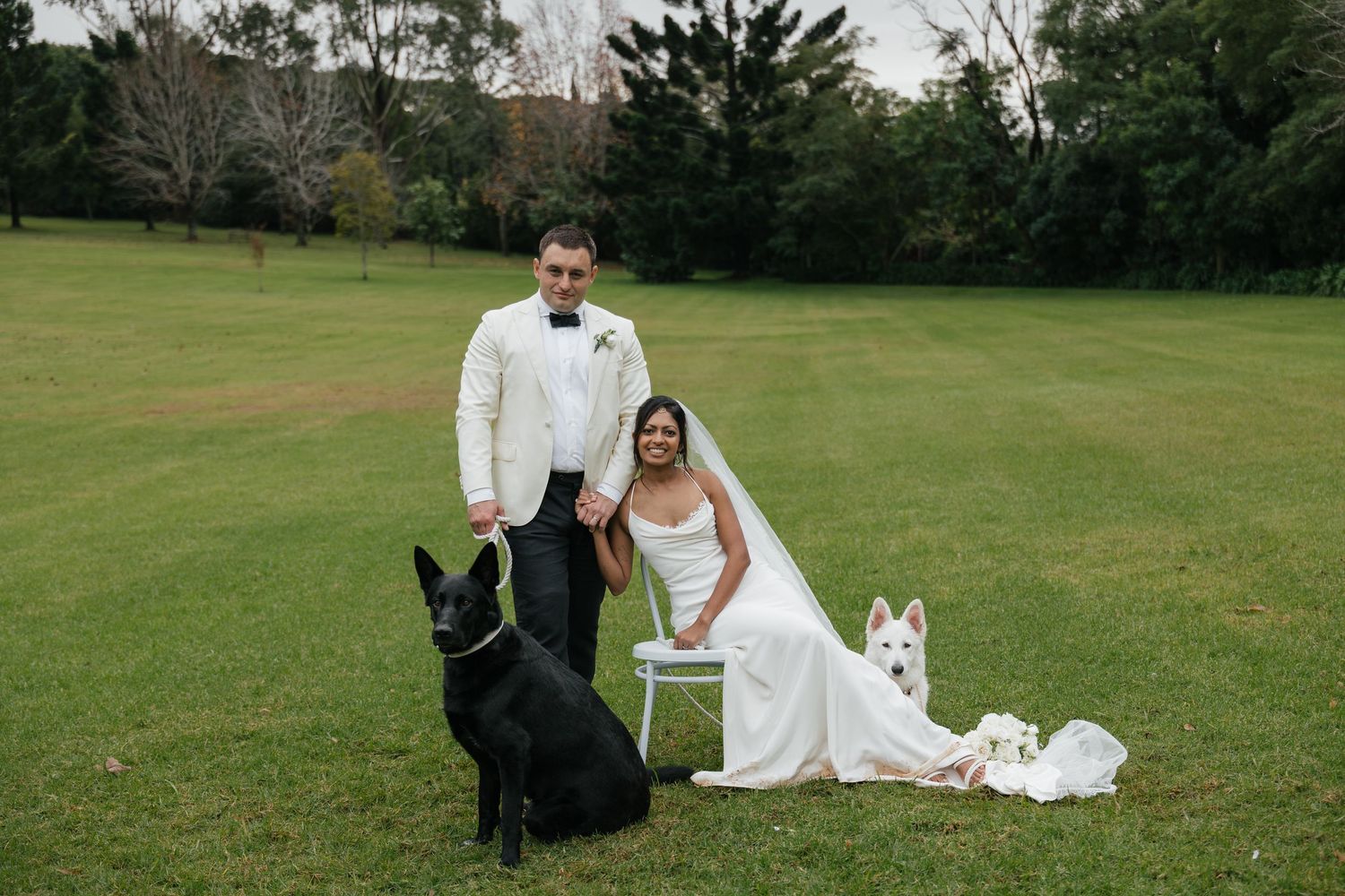 Bride and groom pose with black and white dogs on green lawn in wedding attire outdoors.