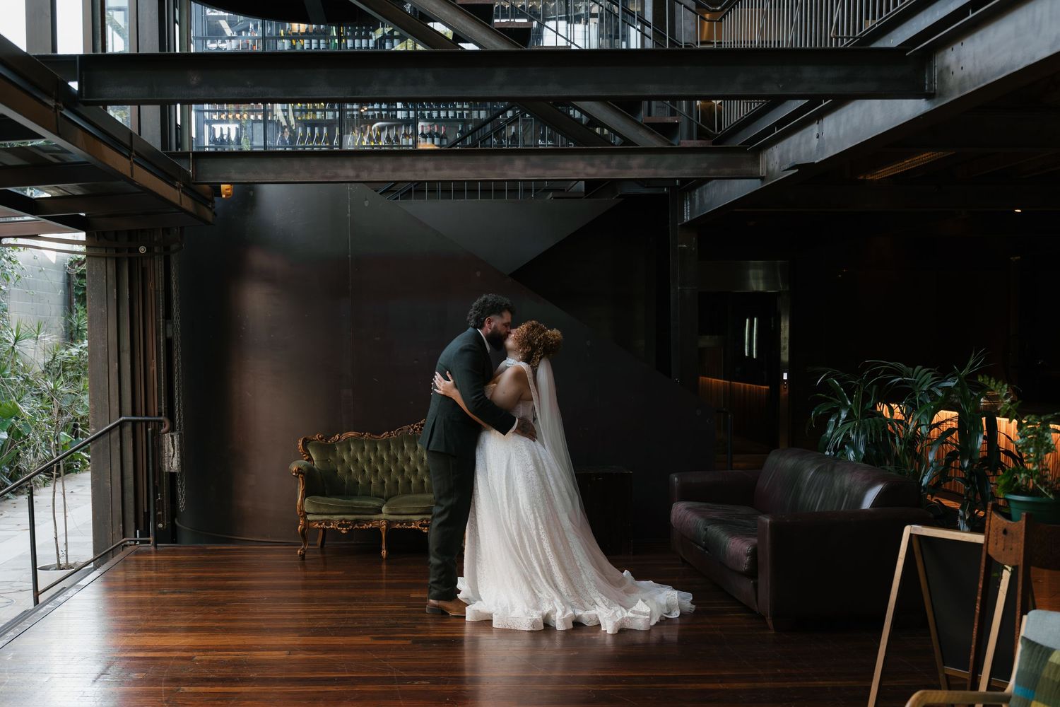 A bride and groom share a romantic embrace in a modern industrial wedding venue with wooden floors and lush greenery.