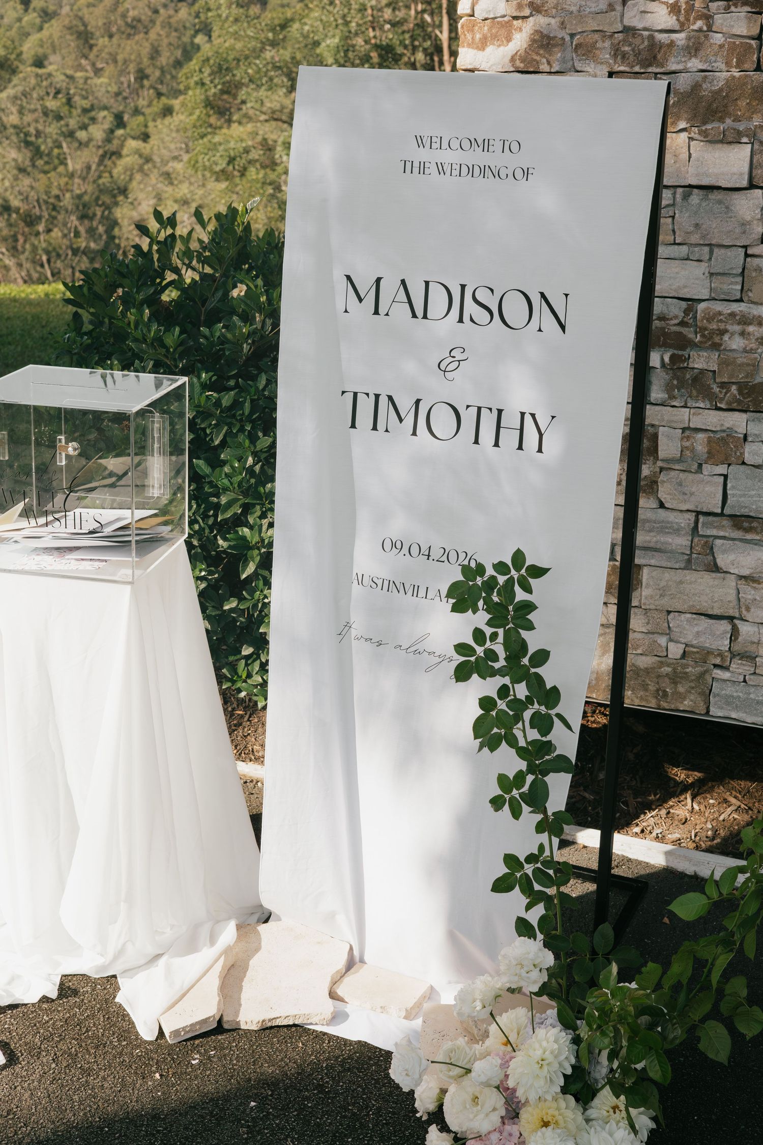 Elegant outdoor wedding welcome sign for Madison and Timothy, adorned with greenery, beside a draped table.
